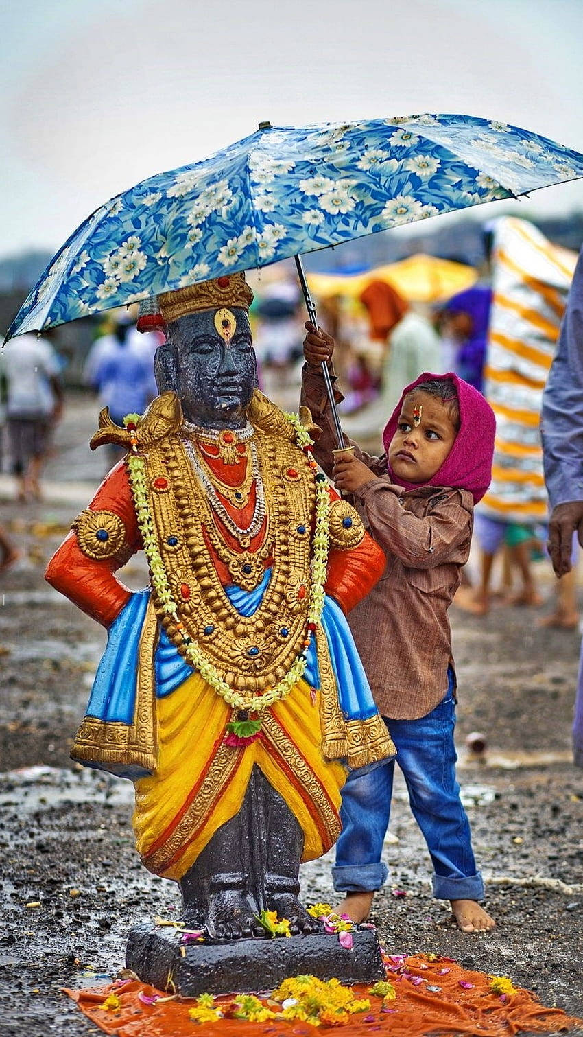 Indian 桌布 壁紙 — 一個穿著粉紅色連帽衫的小孩在雨中節日期間，為一尊色彩繽紛的神像撐著一把花卉圖案的雨傘。