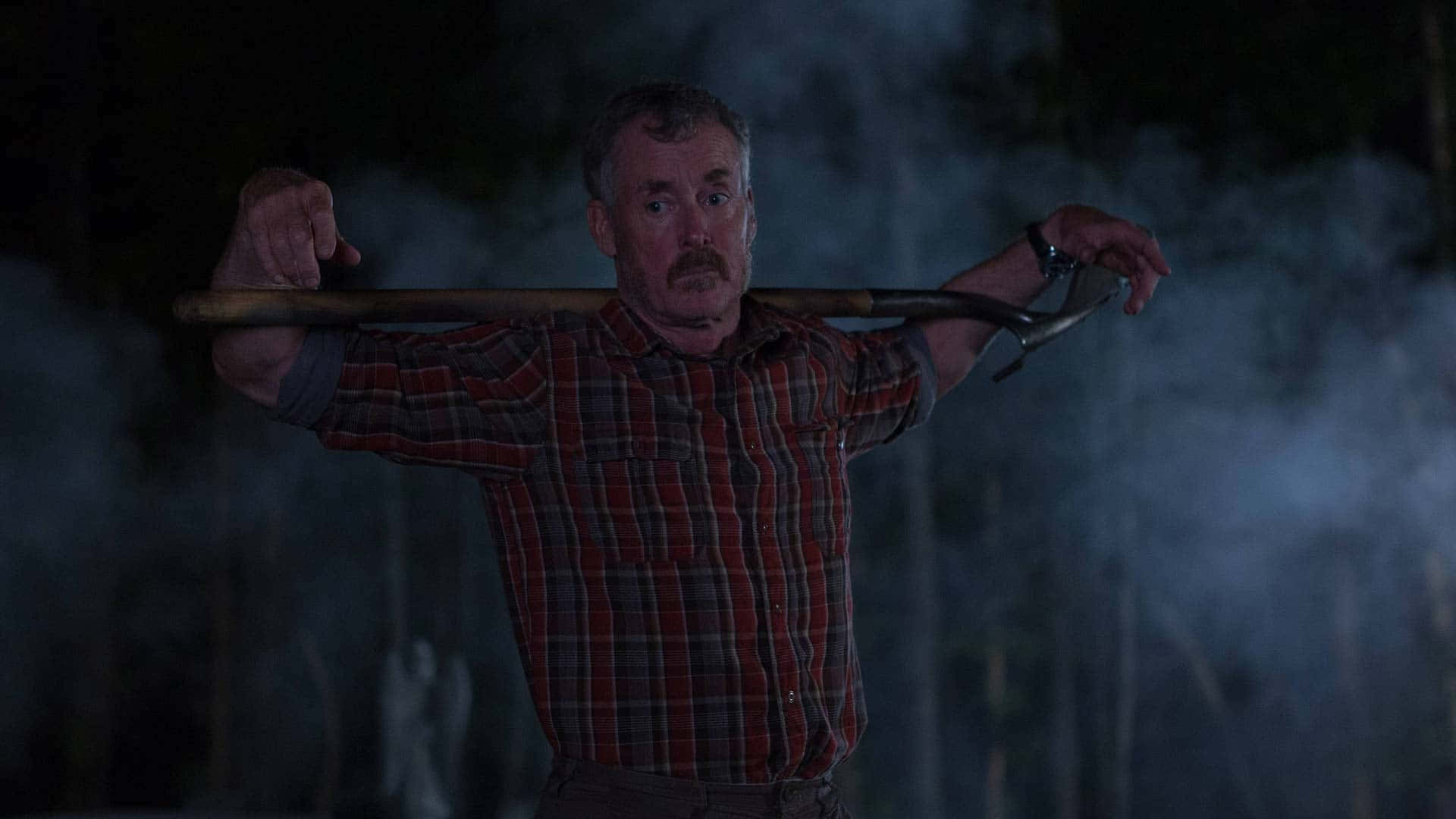 John C. Mcginley 桌布 壁紙 — 一個穿著紅色格仔衫嘅男人喺夜晚煙霧瀰漫嘅黑暗森林中將鏟扛喺肩膀上。