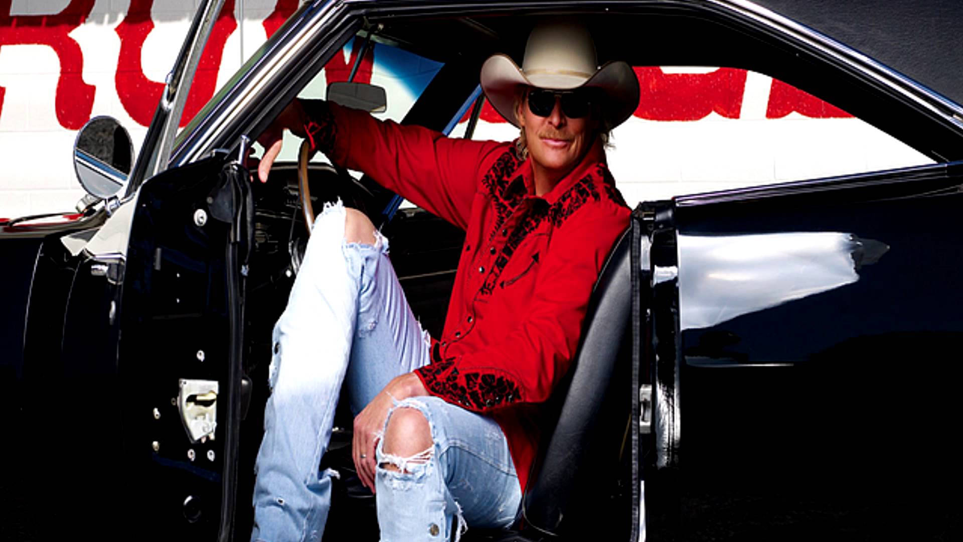 Alan Jackson 桌布 壁紙 — 一個穿著紅色襯衫和破洞牛仔褲的牛仔，自信地笑著從一輛黑色經典汽車中探出身子。