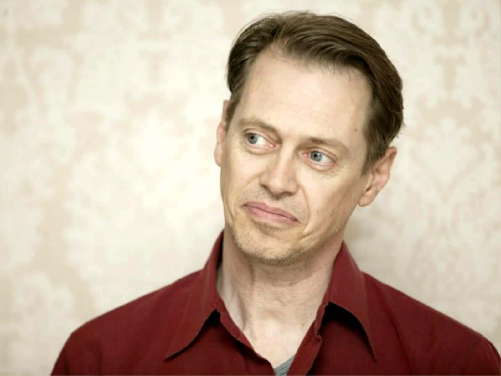steve buscemi 桌布 壁紙 — 一個穿著紅色襯衫的男人，在帶有圖案的米色牆紙背景下，側面露出懷疑的表情。