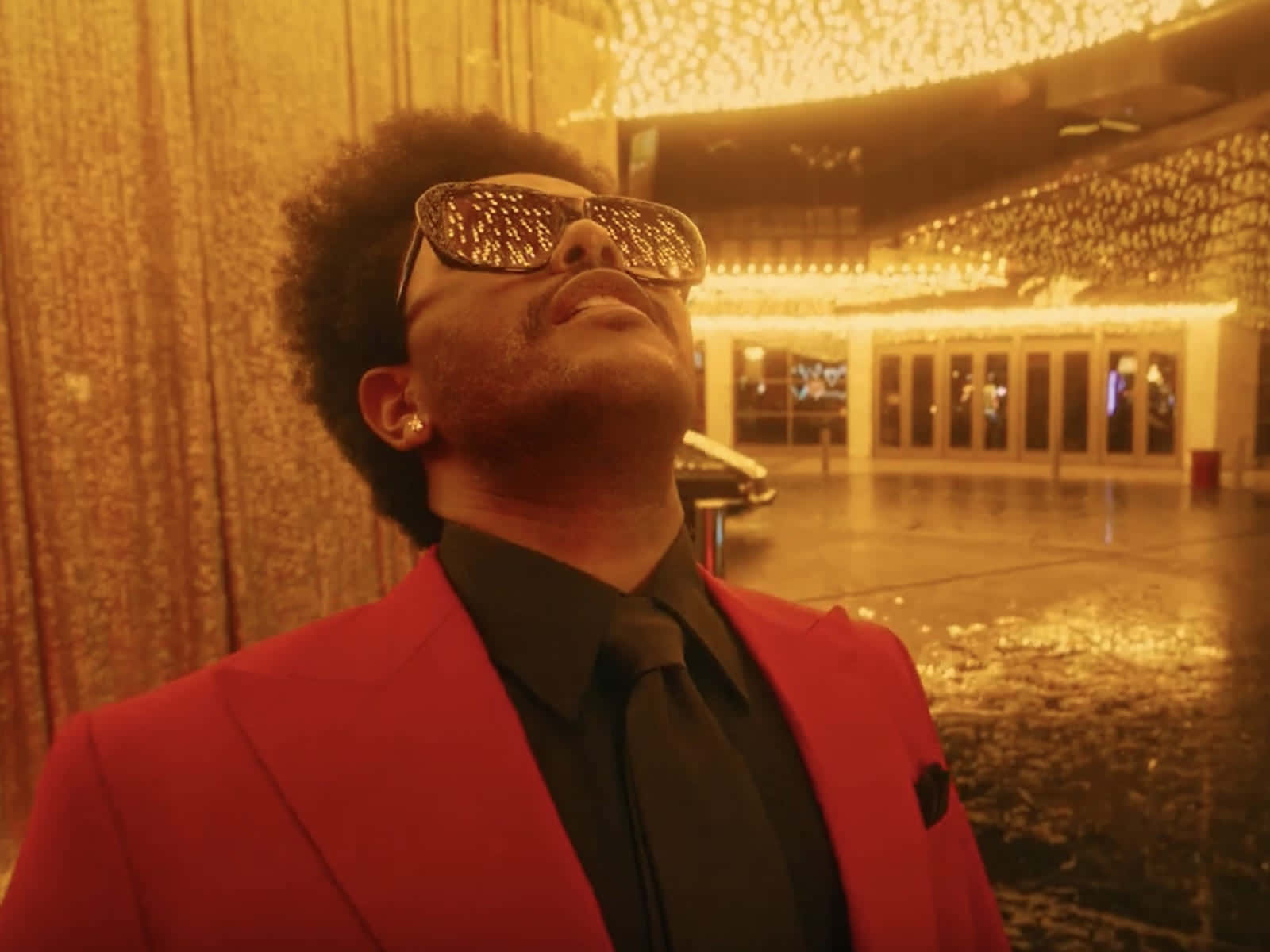 The Weeknd After Hours 桌布 壁紙 — 一個穿著紅色西裝和閃亮太陽眼鏡的男人在潮濕的城市庭院中，在金色燈串下向後仰頭。