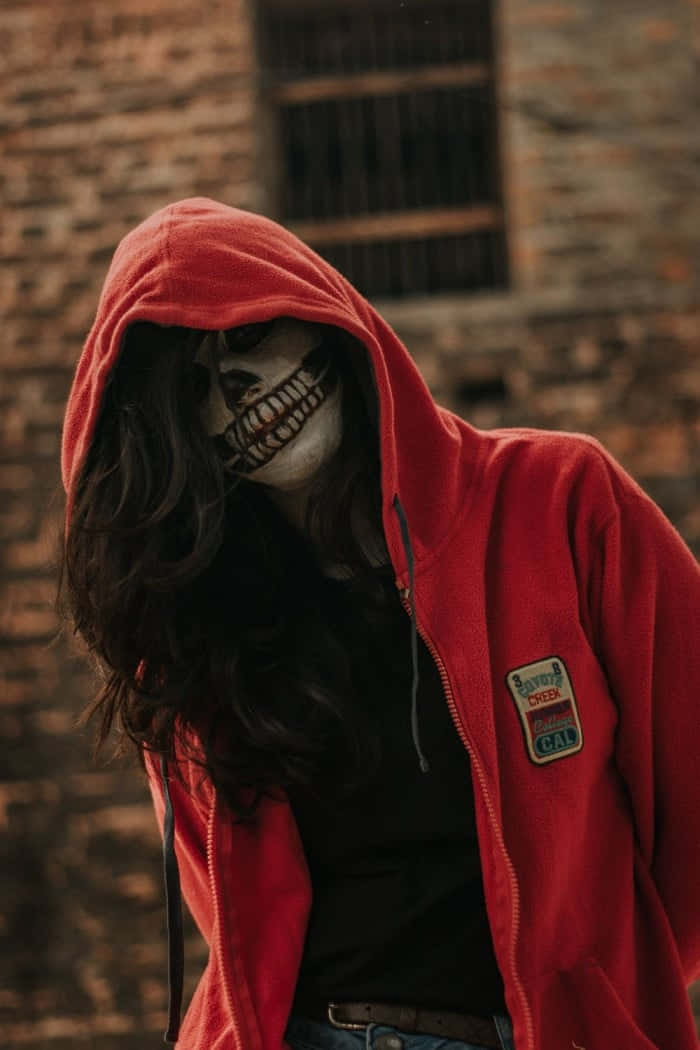 Red Hoodie 桌布 壁紙 — 一個穿著紅色連帽衫和骷髏面具的人站在一堵帶有鐵窗的風化磚牆前。