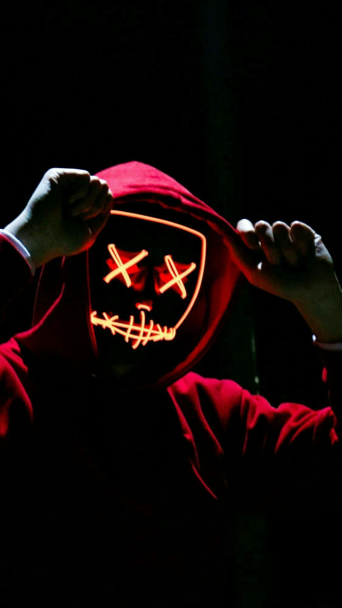 Horror Masks 桌布 壁紙 — 一個穿著紅色連帽衫的人，在黑暗背景前舉起一個發光的橙色LED面具，面具上有X形眼睛和縫合的嘴巴。