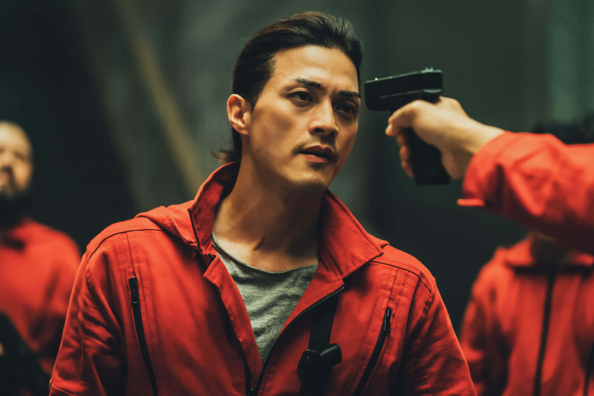 Money Heist 桌布 壁紙 — 一個穿著紅色連身衣的男人在昏暗的房間裡，被一個看不見的人用槍指著頭，他專注地凝視著。