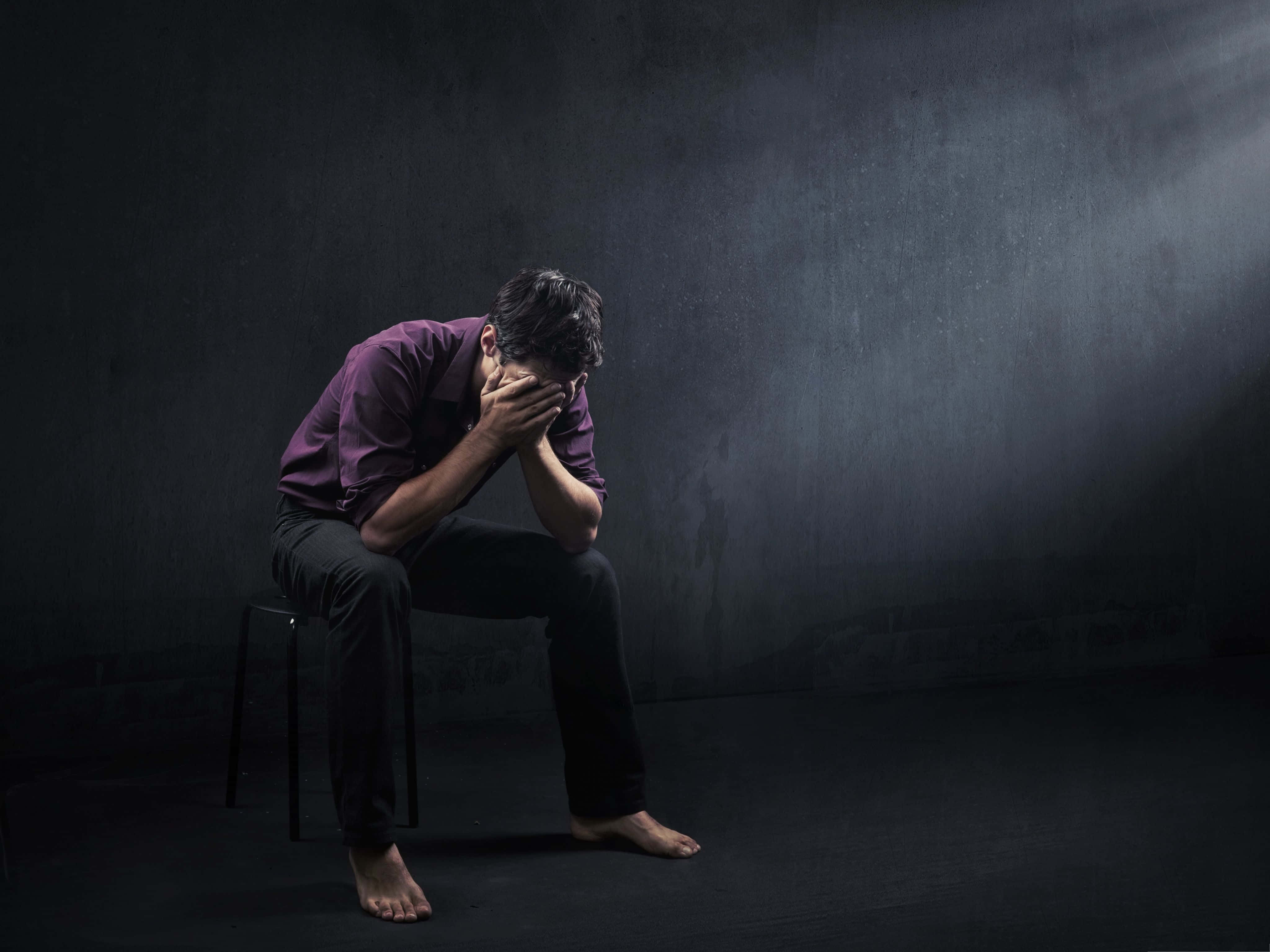 憂鬱 桌布 壁紙 — 一個穿著紫色襯衫的男人獨自坐在昏暗房間的凳子上，絕望地用雙手摀住臉。