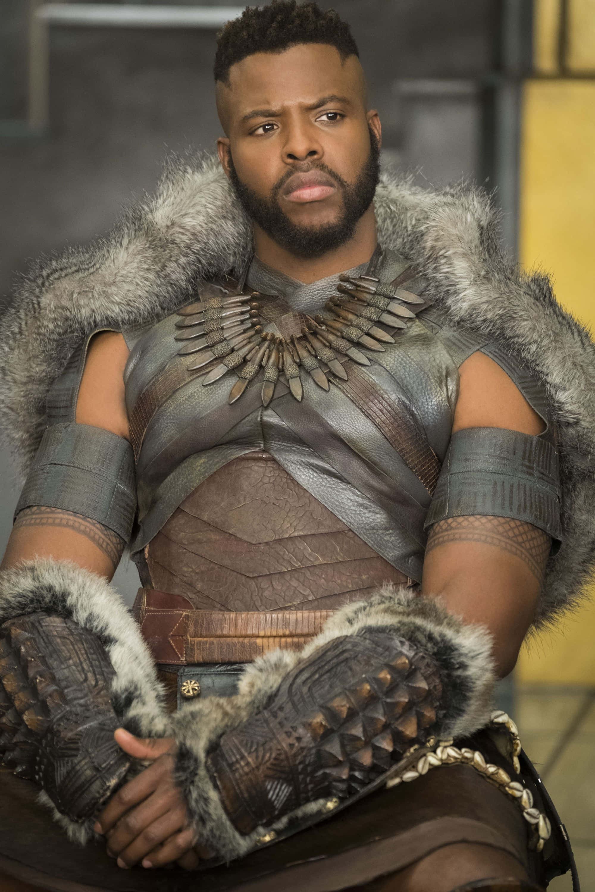 Winston Duke 桌布 壁紙 — 一個穿著細節豐富的瓦干達盔甲、帶有毛皮飾邊和尖刺護手的男人，表情嚴肅地坐著。