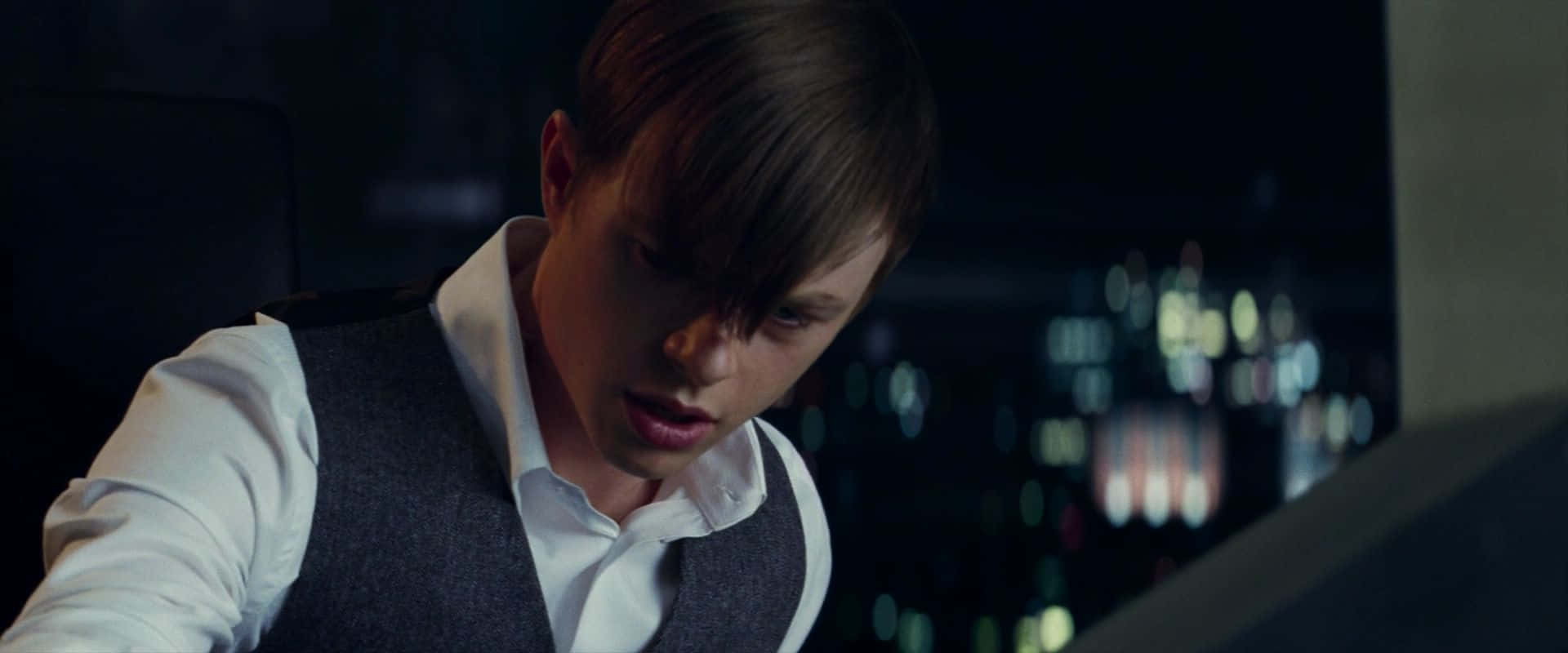 Norman Osborn 桌布 壁紙 — 一個穿著背心的年輕男子在昏暗的房間裏專注地向前傾，身後是模糊的城市燈光。