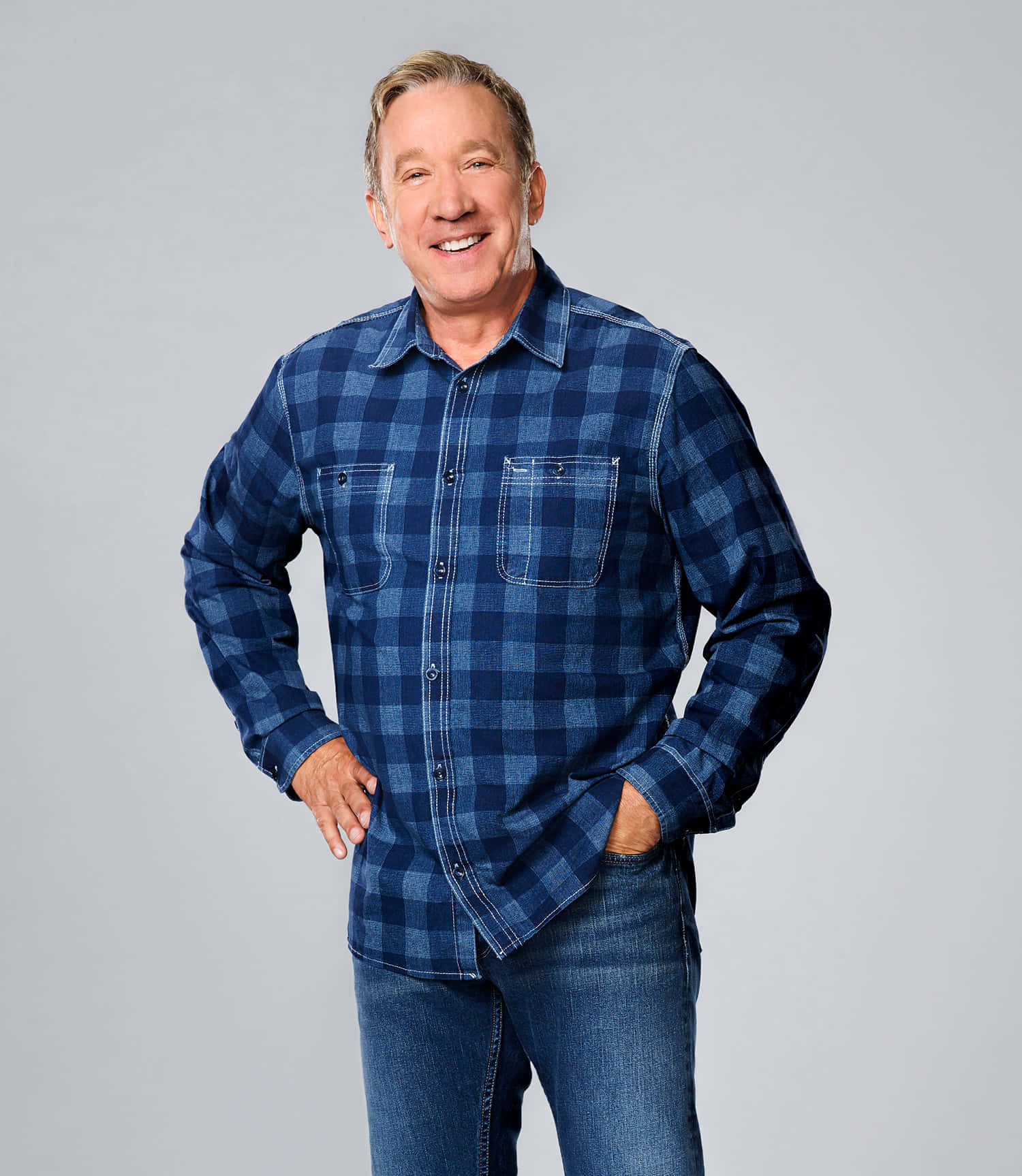 Tim Allen 桌布 壁紙 — 一個穿著藍色格仔恤衫和牛仔褲的開朗男人，雙手插袋，在純灰色背景前微笑。