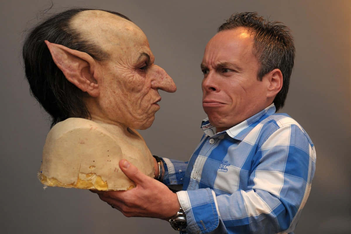 Warwick Davis 桌布 壁紙 — 一個穿著藍色格仔恤衫嘅男人，喺純灰色背景前，拎住一個精細嘅哥布林面具，同時模仿佢誇張嘅面部表情。