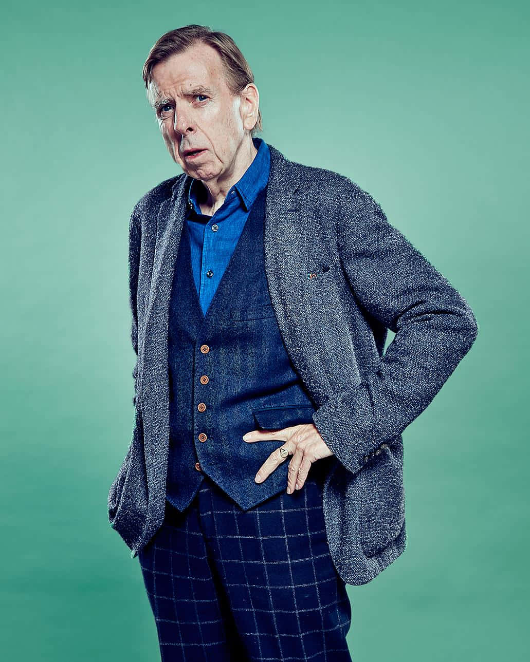 Timothy Spall 桌布 壁紙 — 一個穿著藍色襯衫和粗花呢外套的男人，在純青色背景前擺出懷疑的表情。
