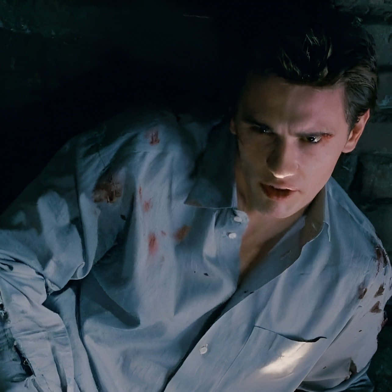Harry Osborn 桌布 壁紙 — 一個穿著血跡襯衫的男人在昏暗的石頭環境中凝視著。
