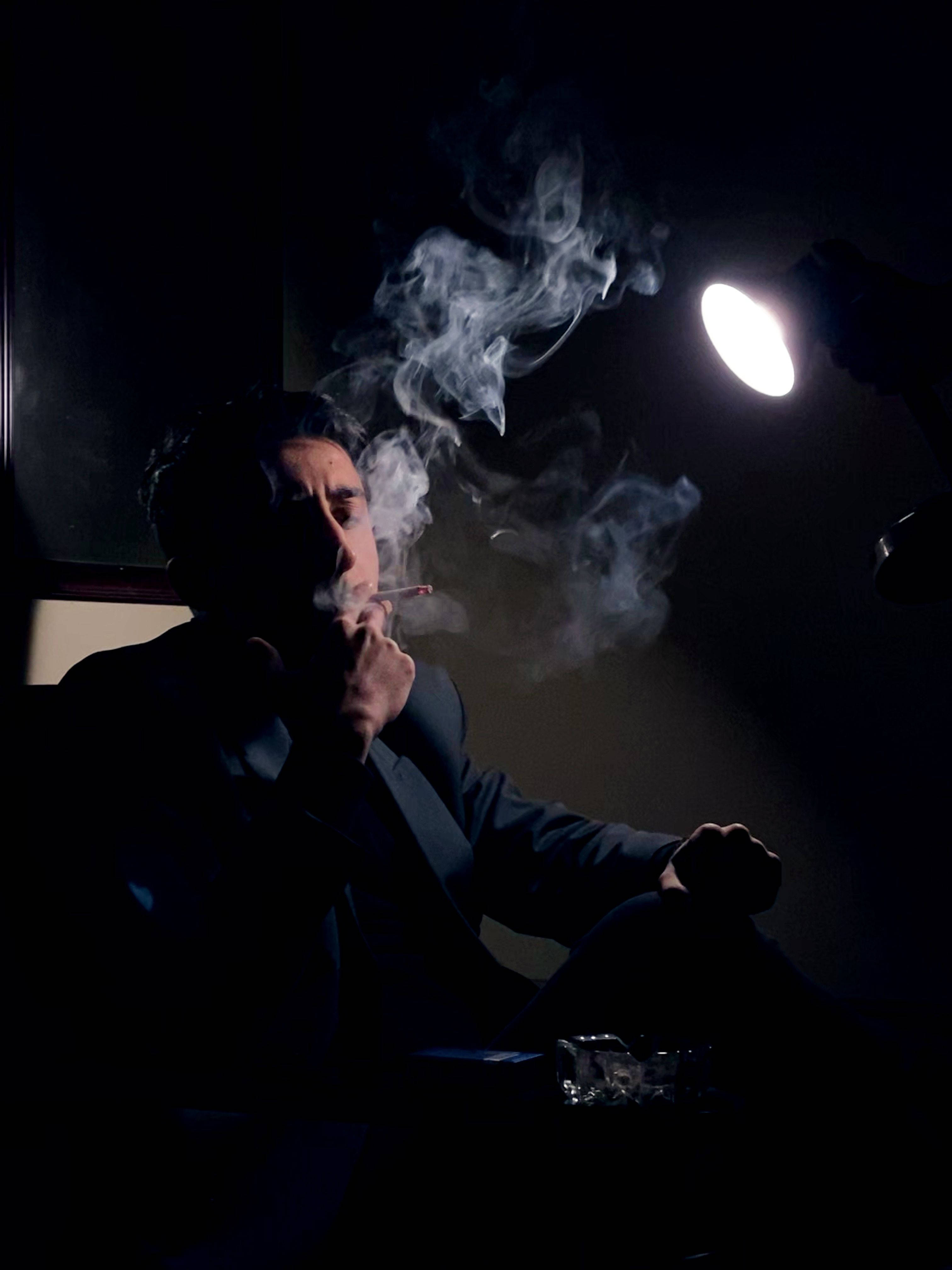 男人 桌布 壁紙 — 一個穿著西裝的男人在黑暗的房間裡，在單一明亮燈光下抽煙，煙霧繚繞。