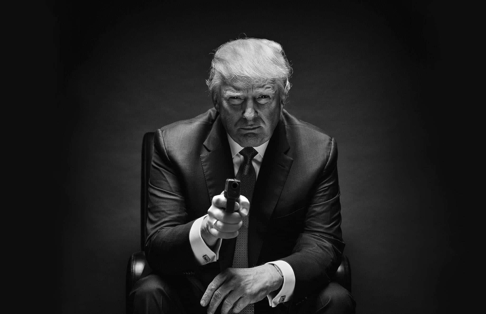 trump 桌布 壁紙 — 一個穿著西裝的男人手持手槍指向前方，表情嚴肅的黑白肖像。
