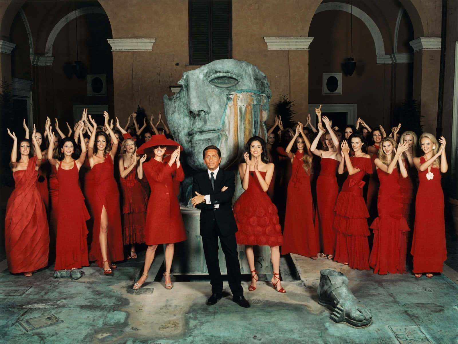 Red Clothing 桌布 壁紙 — 一個穿著西裝的男人站在一個巨大的石臉雕塑前，被穿著鮮豔紅色連衣裙的女人包圍，她們在一個宏偉的庭院中舉起雙手。