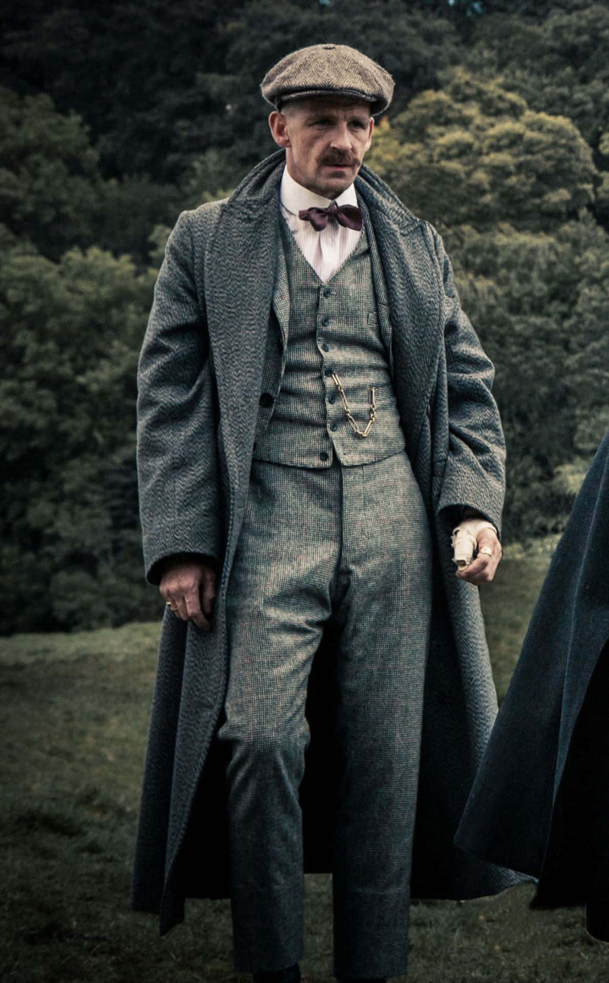 Peaky Blinders 桌布 壁紙 — 一個穿著質感灰色三件套西裝和扁平帽的男人莊嚴地站在樹林田野中，手持一把手槍。