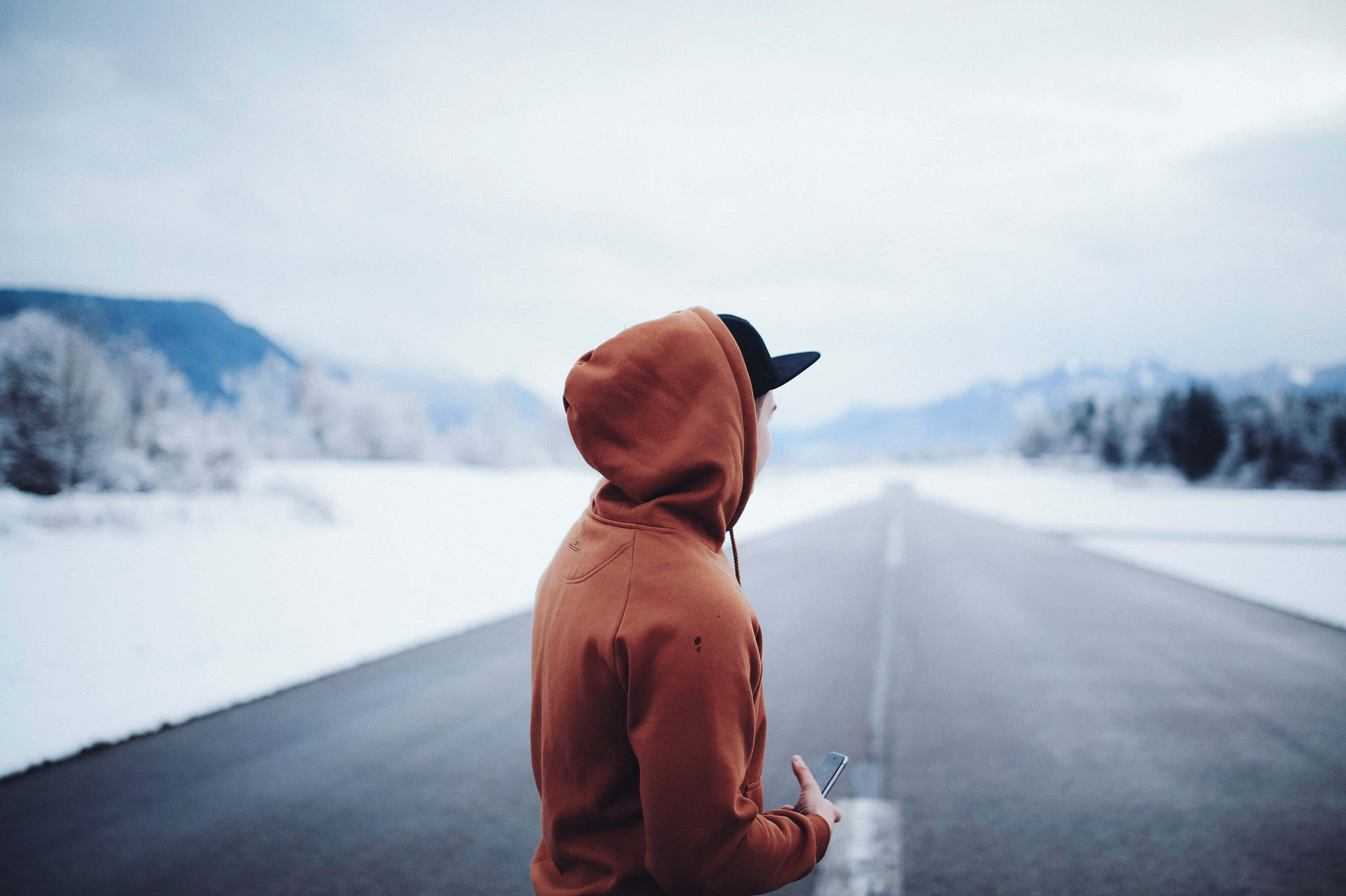 寒冷 桌布 壁紙 — 一個穿著鏽色連帽衫的人獨自站在積雪的道路上，凝視著陰天天空下遠處積雪的山峰。