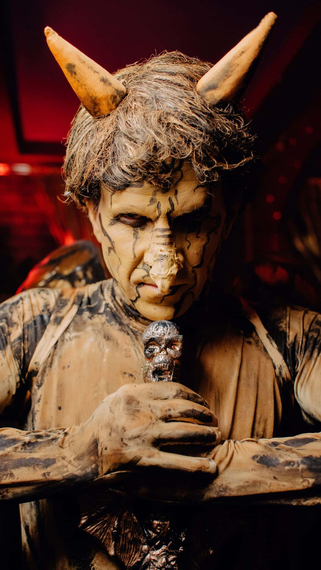 Horror Costumes 桌布 壁紙 — 一個穿著魔鬼服裝、頭上有角、臉上塗有裂紋化妝的人，在戲劇性的紅光下拿著一個頭骨。