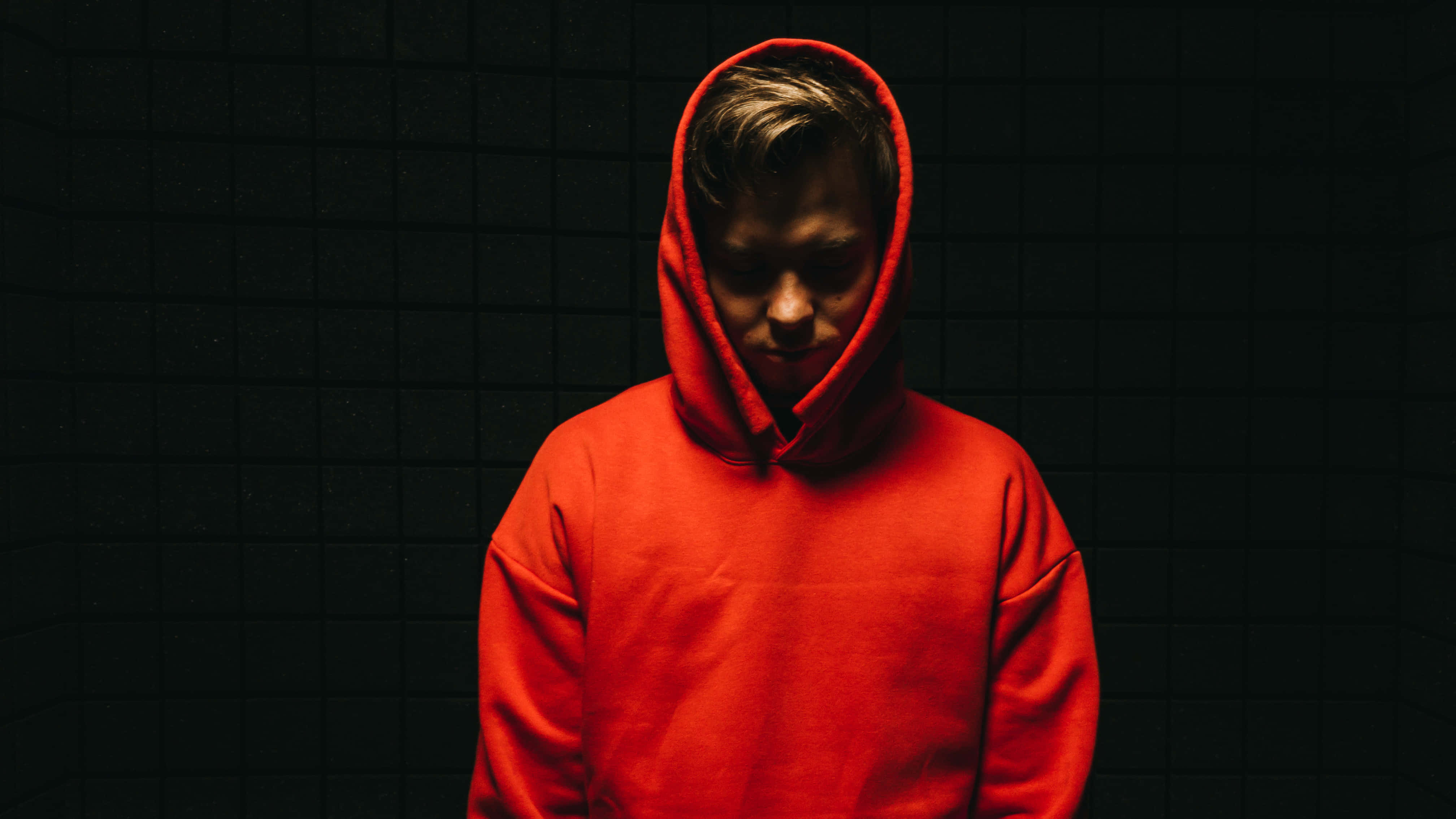 Red Hoodie 桌布 壁紙 — 一個穿著鮮紅色連帽衫的人站在深色瓷磚牆前，低調的燈光在他們的臉上投下陰影。