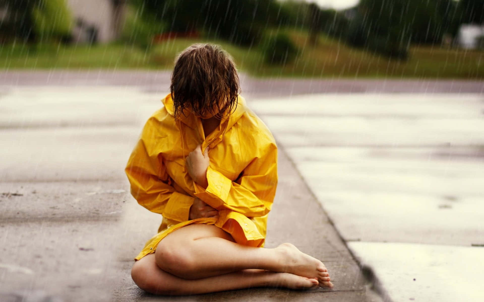 Yellow Raincoat 桌布 壁紙 — 一個穿著鮮黃色雨衣的孩子蜷縮在濕漉漉的人行道上，低著頭，雨水在他們周圍落下。