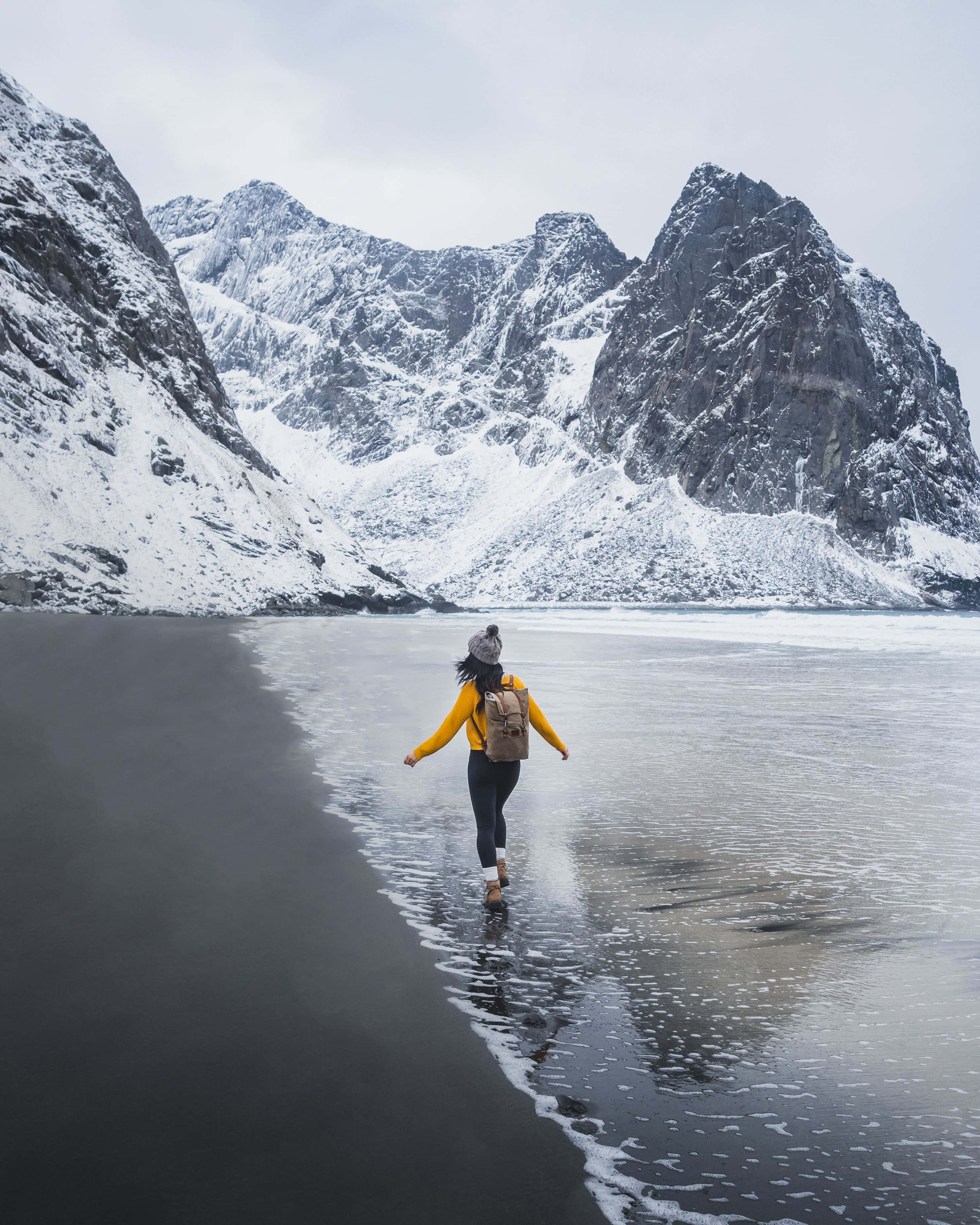 Winter Hiking 桌布 壁紙 — 一個穿著黃色外套的人沿著黑色火山海灘行走，背景是白雪覆蓋的山脈。