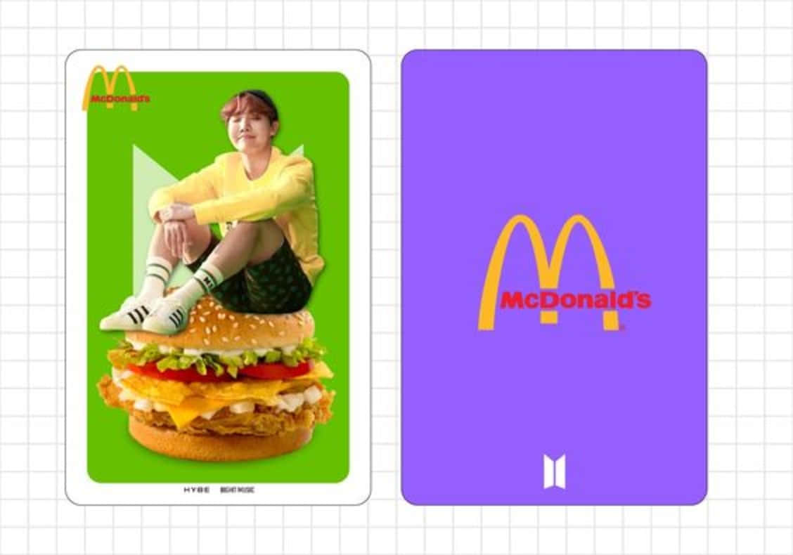 BTS Mac Hobi 壁紙 — 一個穿著黃色連帽衫的人坐在綠色卡片上的一個巨型漢堡上，旁邊是一張帶有BTS標誌的紫色麥當勞卡片。