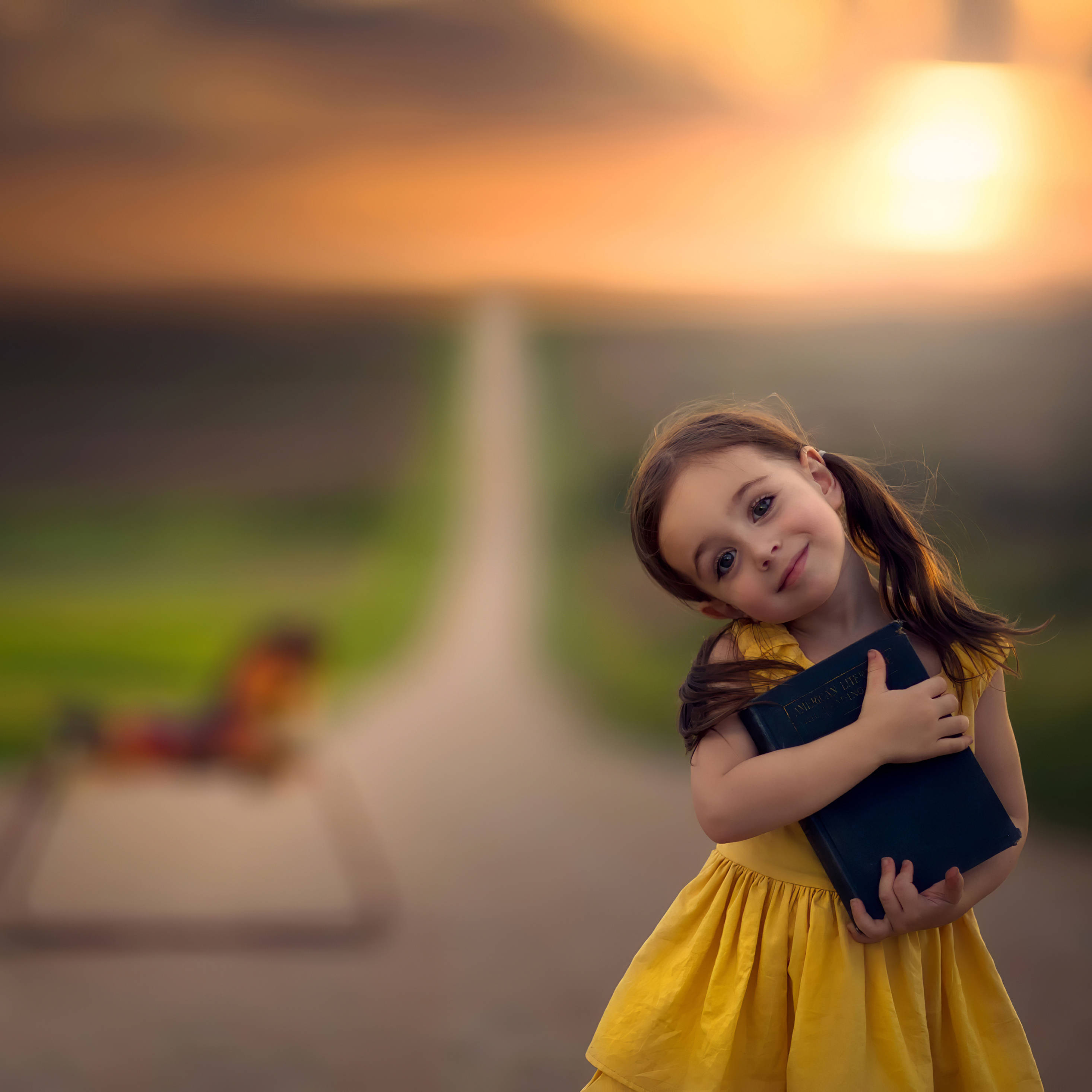 girls 桌布 壁紙 — 一個穿著黃色連衣裙的小女孩在日落時分的路上抱著一本書，背景模糊。