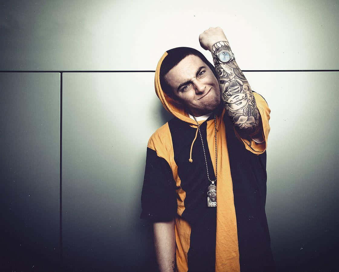 Mac Miller 桌布 壁紙 — 一個穿著黑色和橙色連帽衫、手臂有紋身的男人對著一面純灰色的牆壁繃緊肌肉。