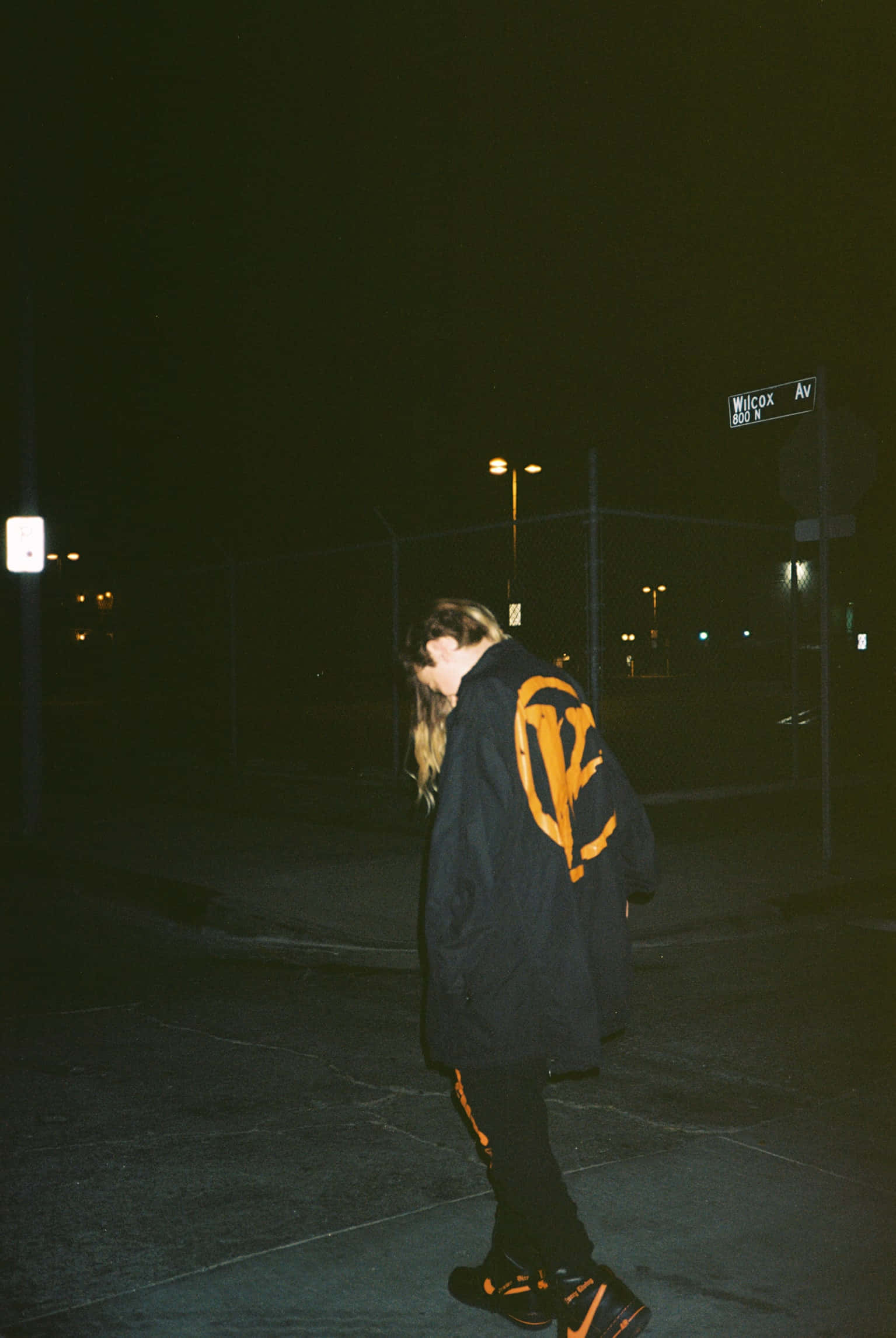 Vlone 桌布 壁紙 — 一個穿著黑色外套、帶有醒目橙色標誌的人，在夜晚獨自走在寧靜的城市街道的路燈下。