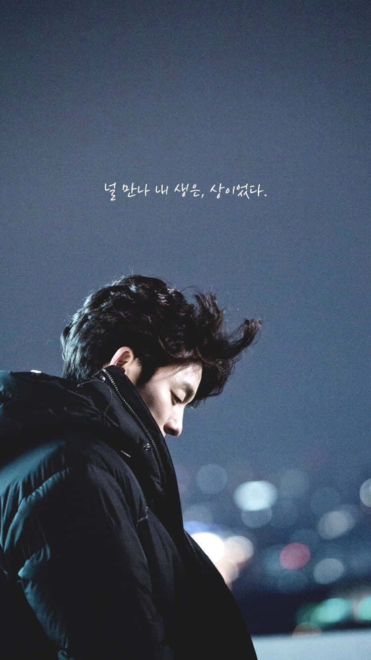 Aesthetic Korean 桌布 壁紙 — 一個穿著黑色外套的年輕男子，在模糊的城市夜景背景中，伴隨著柔和散景燈光向下凝視。