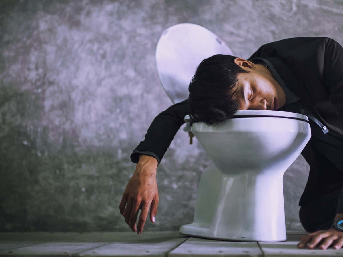 醉咗 桌布 壁紙 — 一個穿著黑色外套的男人在一個牆壁破裂、地板鋪瓷磚的骯髒浴室裡對著馬桶嘔吐。
