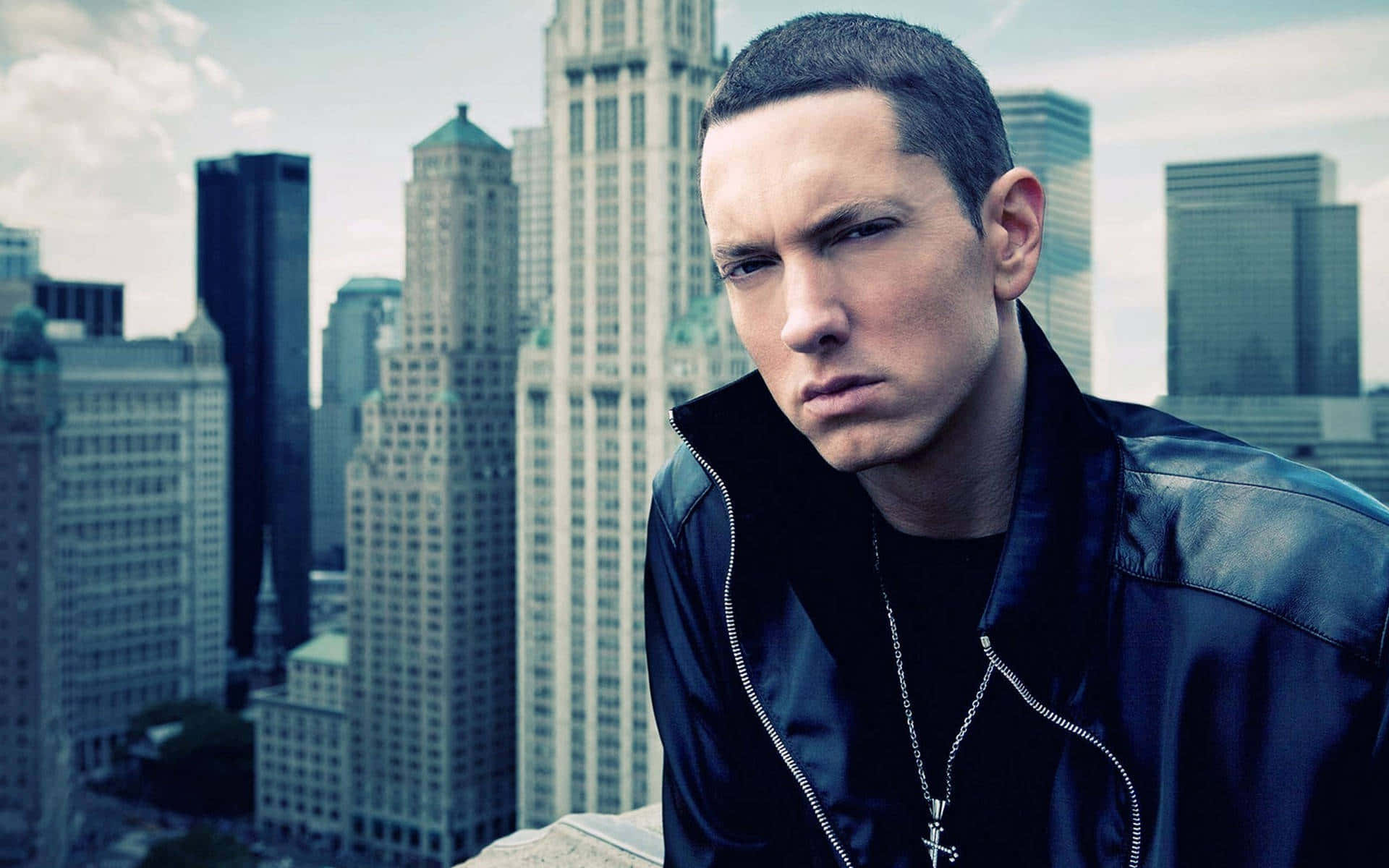 Eminem 桌布 壁紙 — 一個穿著黑色皮褸的男人，在陰天高聳的城市摩天大樓背景下，眼神銳利地凝視著。