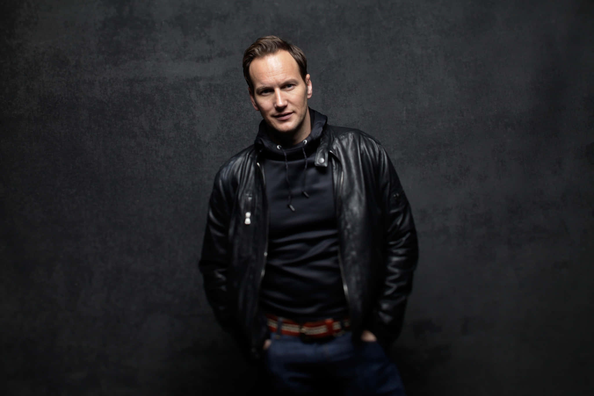 Patrick Wilson 桌布 壁紙 — 一個穿著黑色皮褸的男人，雙手插袋，自信地站在有紋理的深灰色攝影棚背景前。