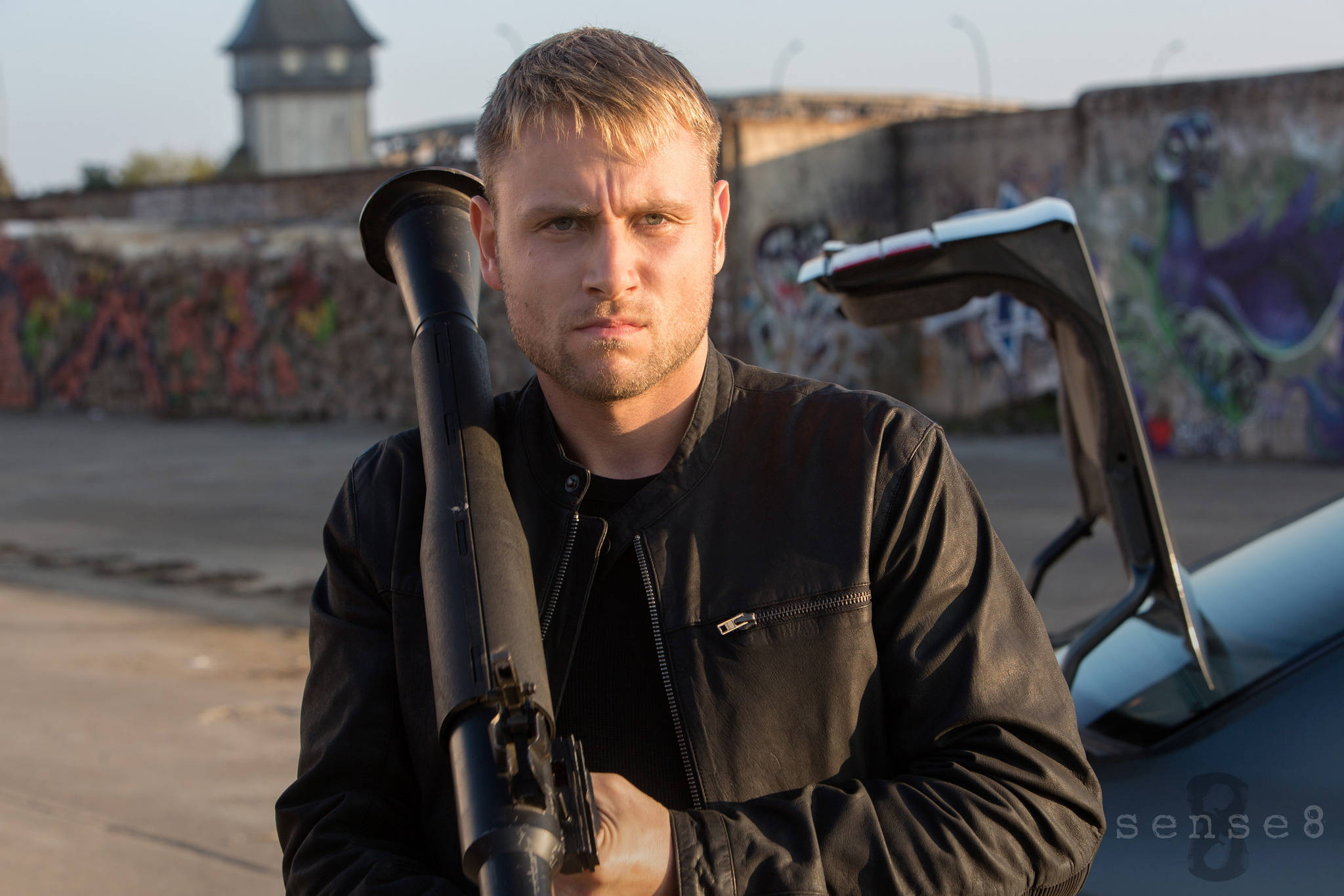 《Sense8》Wolfgang Bogdanow 壁紙 — 一個穿著黑色皮褸的男人喺一架打開車門嘅車旁邊，對住一幅佈滿塗鴉嘅牆，揸住一支火箭炮。