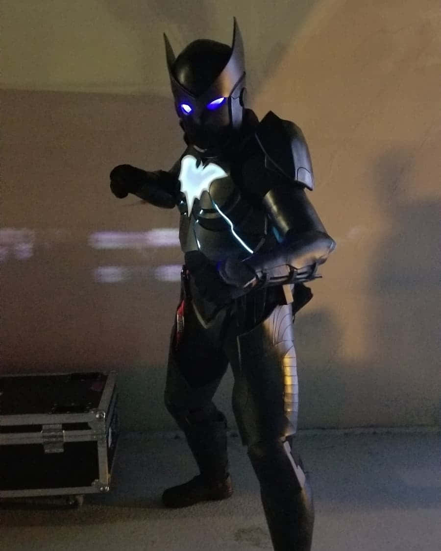 Batwing 桌布 壁紙 — 一個穿著黑色裝甲蝙蝠俠服裝、擁有發光藍色眼睛和胸部標誌的人站在燈光昏暗的房間裡。