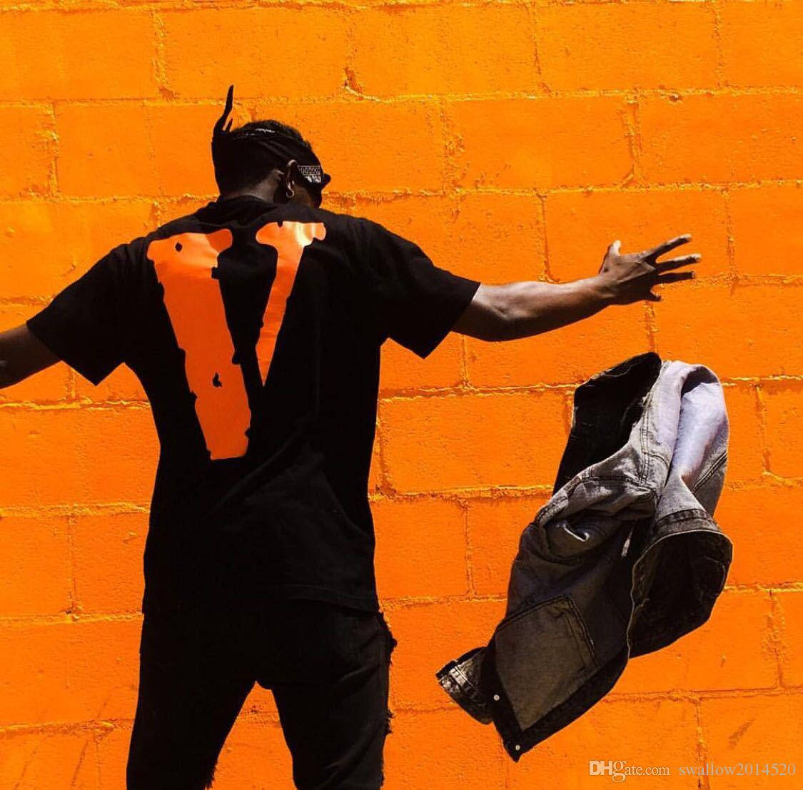 Vlone 桌布 壁紙 — 一個穿著黑色襯衫、背後有橙色「V」字的人，站在充滿活力的橙色磚牆前，手持一件牛仔外套。