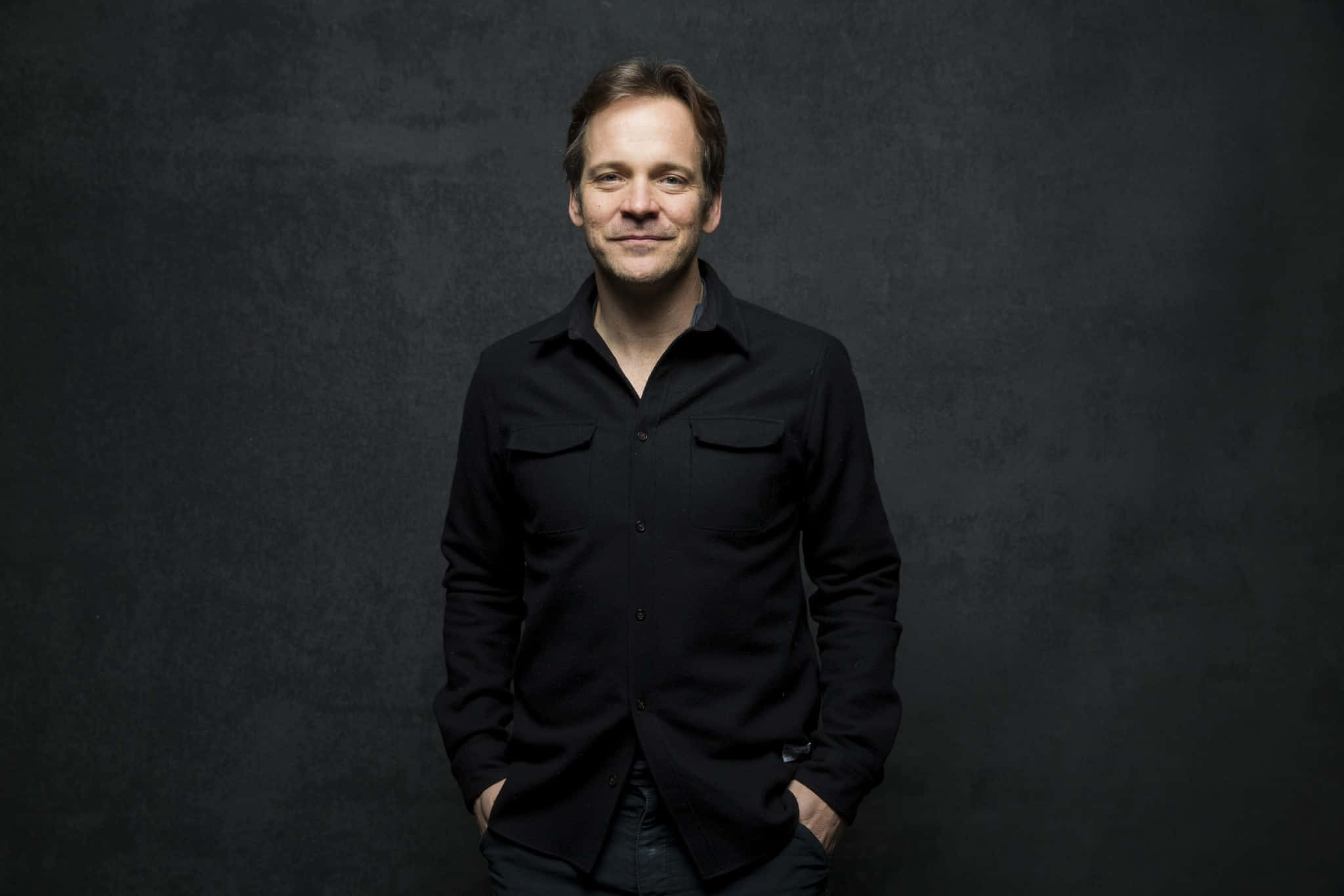 Peter Sarsgaard 桌布 壁紙 — 一個穿著黑色襯衫的男人自信地站在有紋理的深灰色背景前，雙手插在口袋裡。
