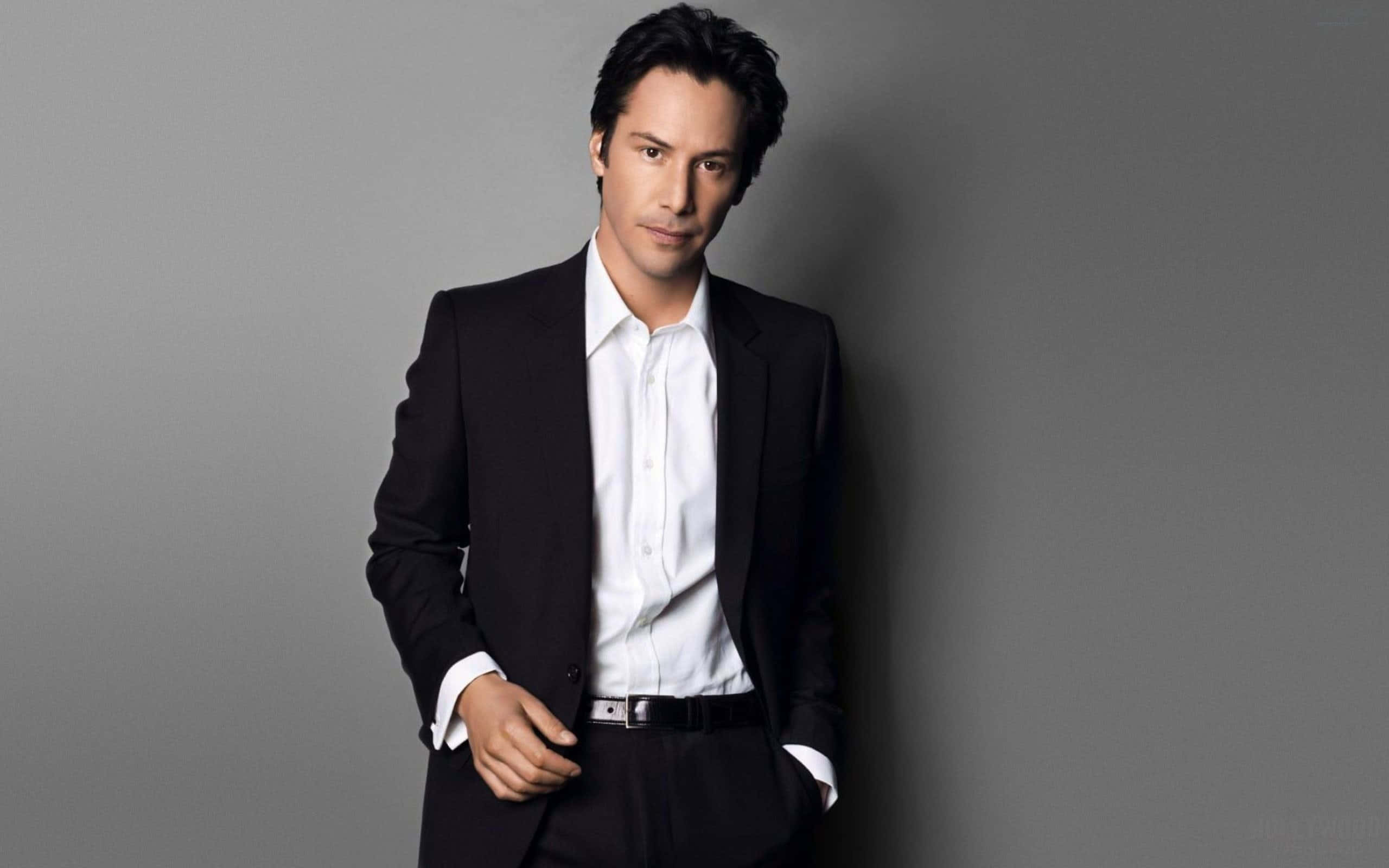 Keanu Reeves 桌布 壁紙 — 一個穿著黑色西裝和白色襯衫的男人，一隻手插在口袋裡，自信地靠著一面純灰色的牆。