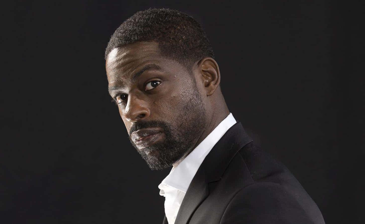 Sterling K Brown 桌布 壁紙 — 一個穿著黑色西裝和白色襯衫的男人在黑暗背景下強烈地凝視。