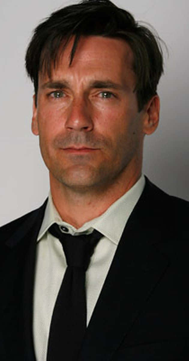 Jon Hamm 桌布 壁紙 — 一個穿著黑色西裝和領帶的男人，在純灰色背景下，表情嚴肅地直視鏡頭。