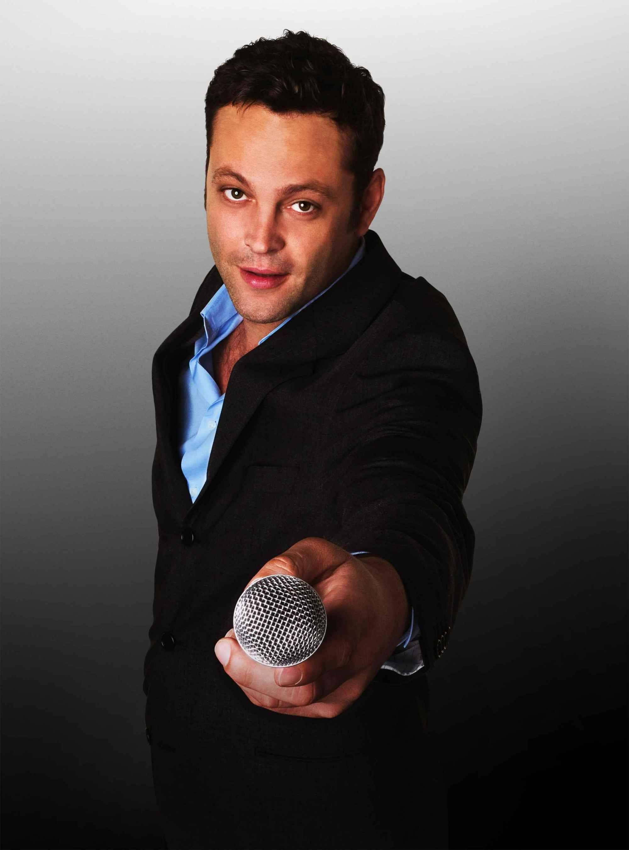 Vince Vaughn 桌布 壁紙 — 一個穿著黑色西裝外套和藍色襯衫的男人，在灰色漸變背景前，將麥克風朝向觀眾。