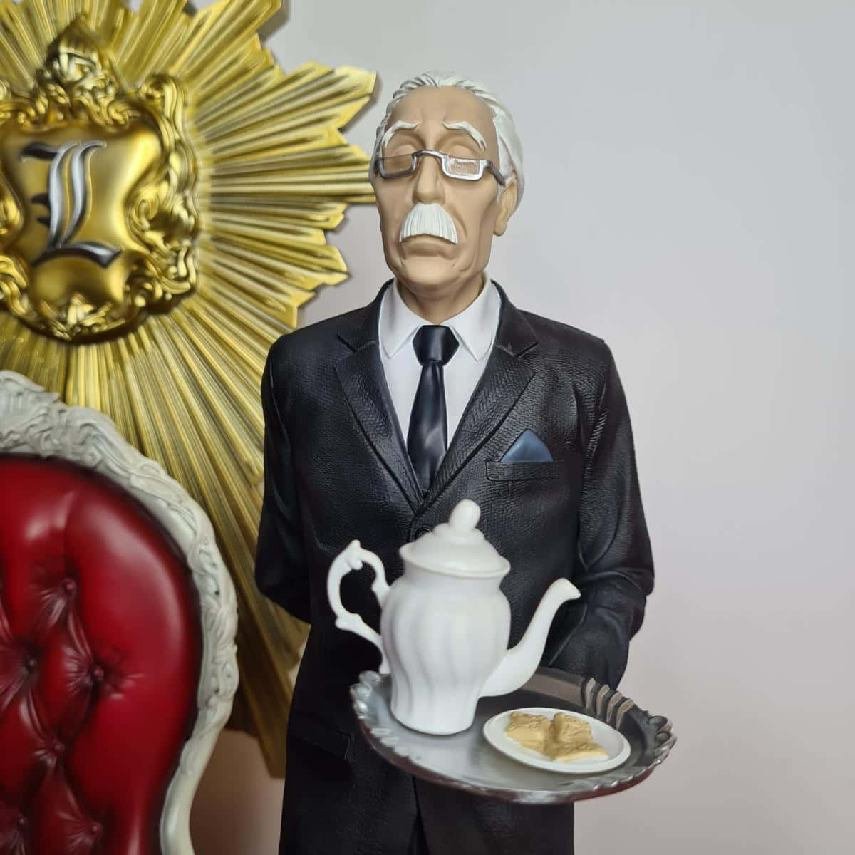 Death Note Watari 桌布 壁紙 — 一個穿著黑色西裝的老年男子手持茶具的詳細雕像，旁邊有一張紅色簇絨椅子和一個金色太陽光芒標誌。