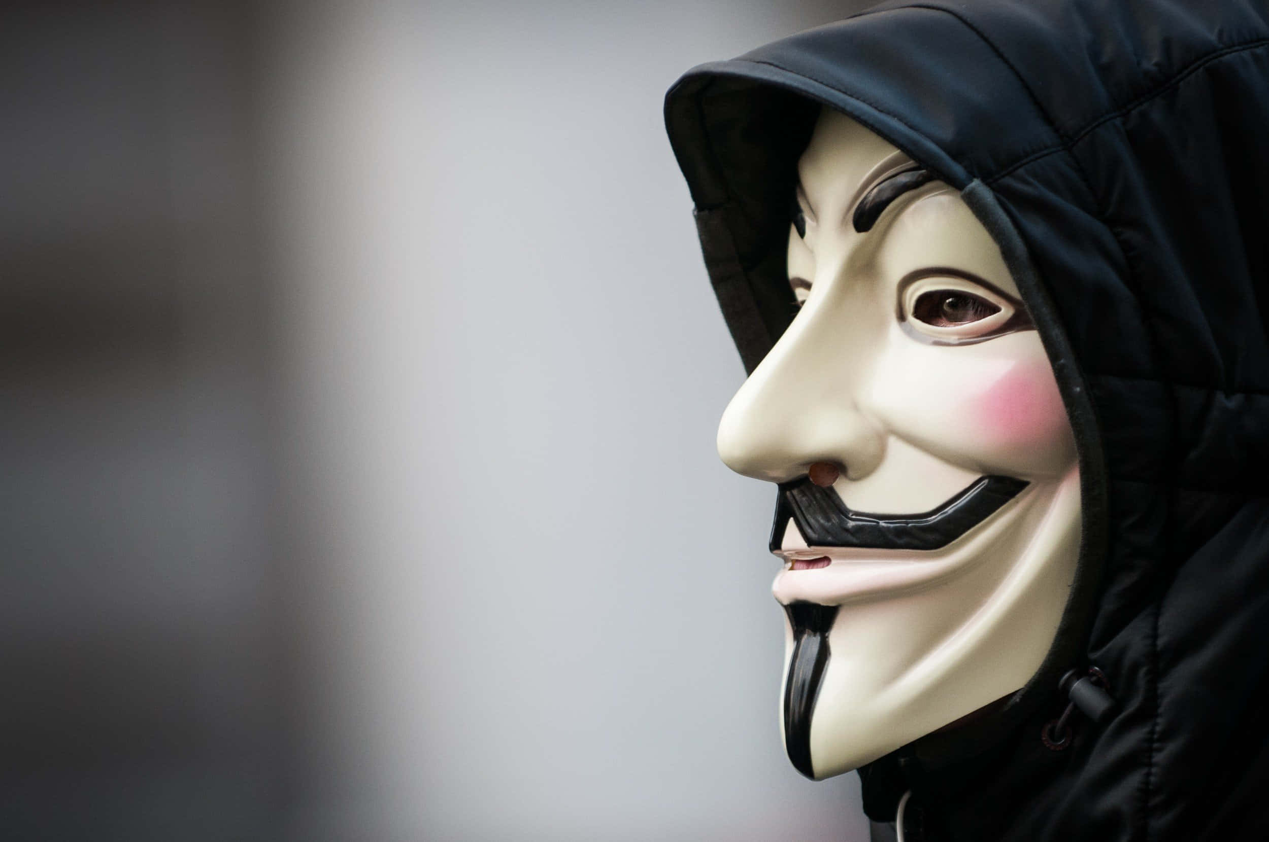 Anonymous 桌布 壁紙 — 一個穿著黑色連帽外套的人，在模糊的城市背景中戴著白色蓋伊·福克斯面具，面具上有寬闊的笑容和粉紅色的臉頰。