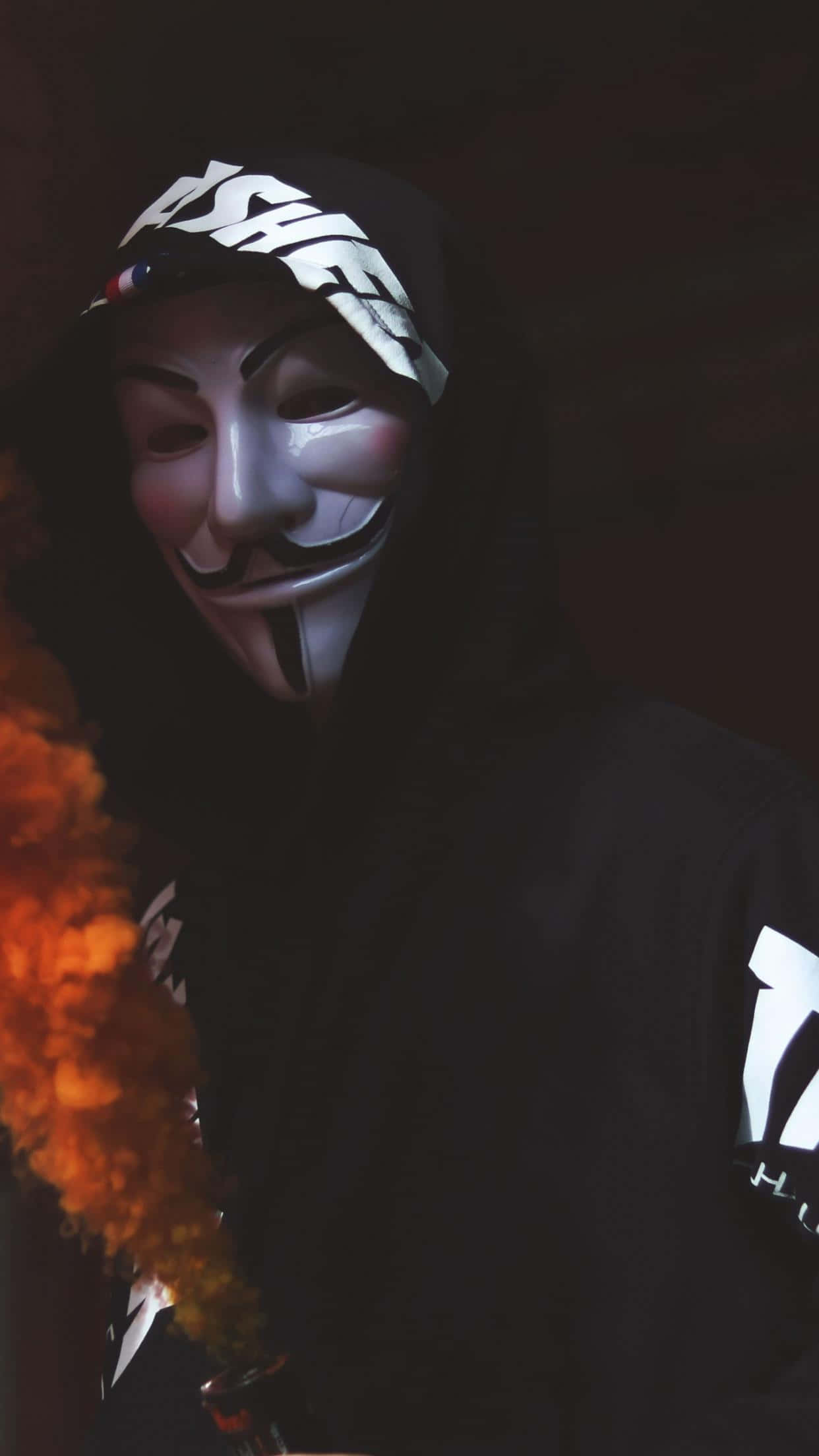 Anonymous 桌布 壁紙 — 一個穿著黑色連帽衫、戴著白色蓋伊·福克斯面具的人，在黑暗背景下呼出橙色煙霧。