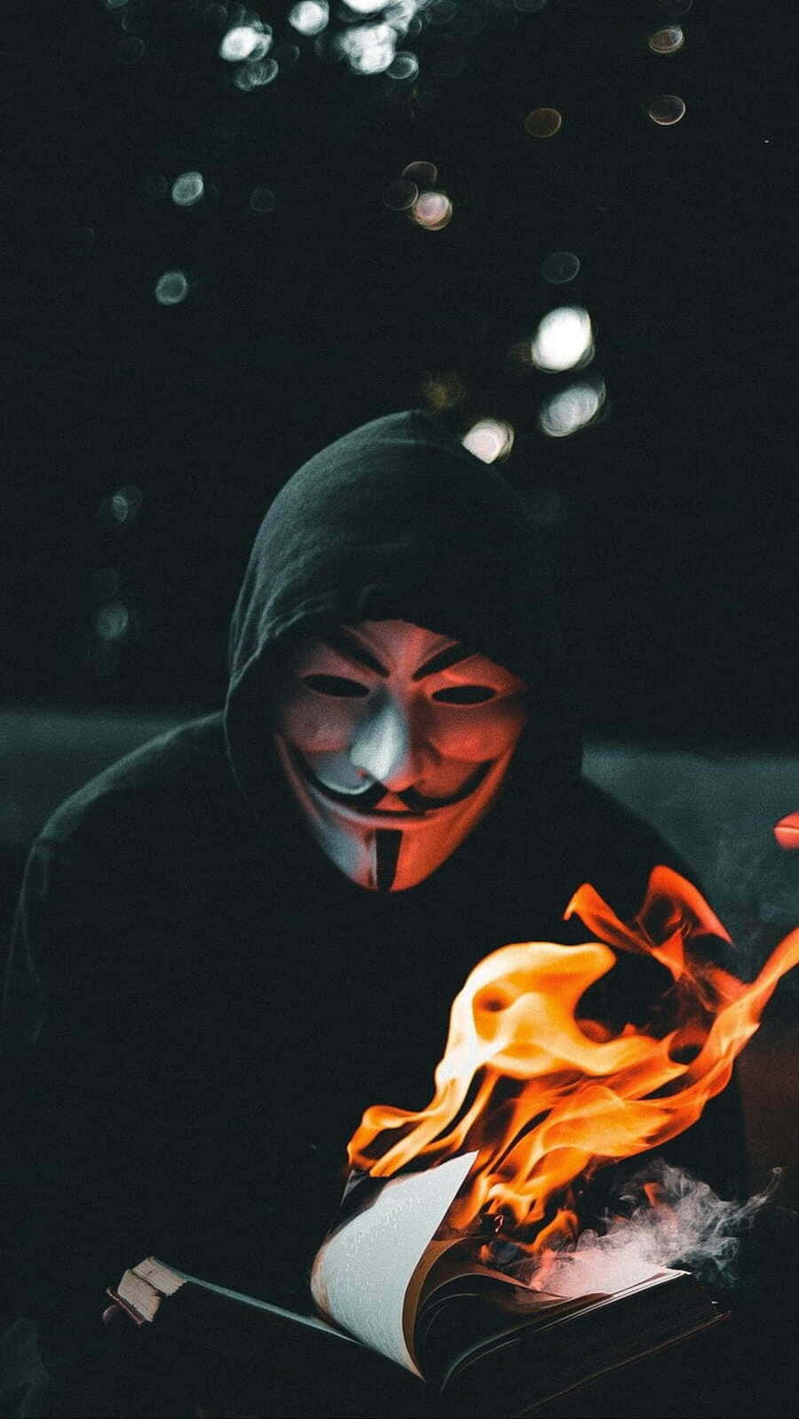  壁紙 — 一個穿著黑色連帽衫、戴著蓋伊·福克斯面具的人，在黑暗背景中閃爍的散景燈光下，手持一本被明亮橙色火焰吞噬的書。