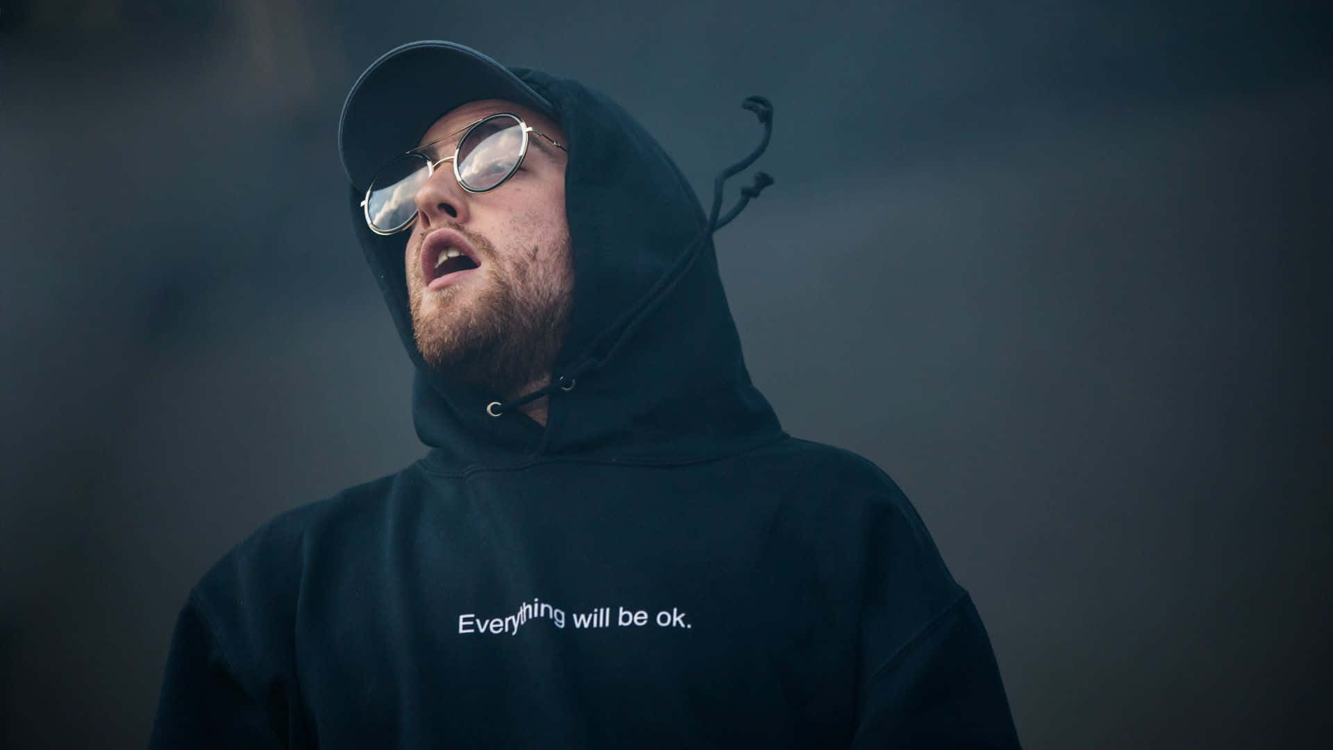 Mac Miller 桌布 壁紙 — 一個穿著黑色連帽衫和圓形太陽眼鏡的男人，張開嘴巴向上看，背景是煙霧瀰漫的黑暗。