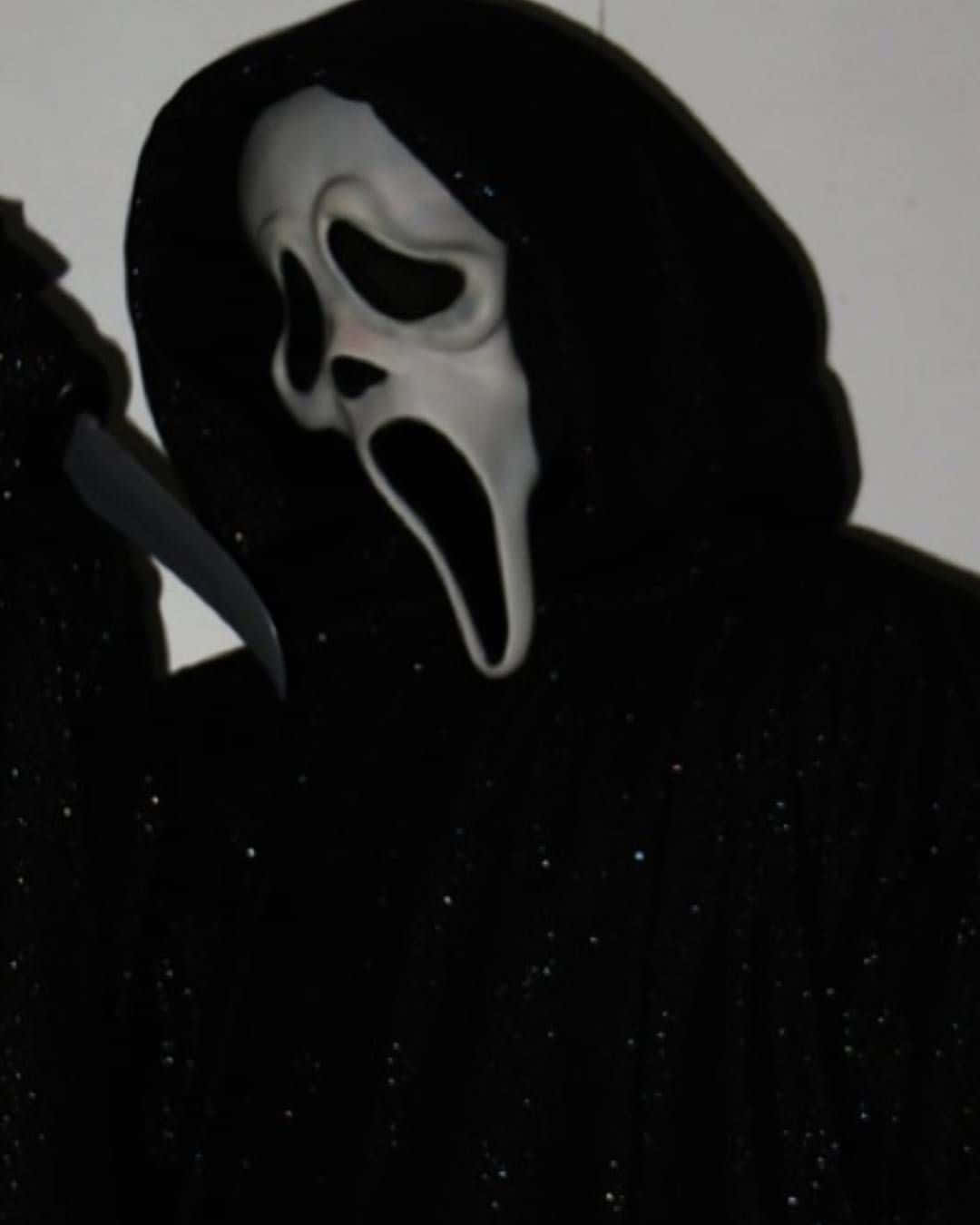 Ghostface Aesthetic 桌布 壁紙 — 一個穿著黑色連帽長袍、戴著標誌性白色驚聲尖叫面具的威脅人物，拿著刀靠著一面素牆。