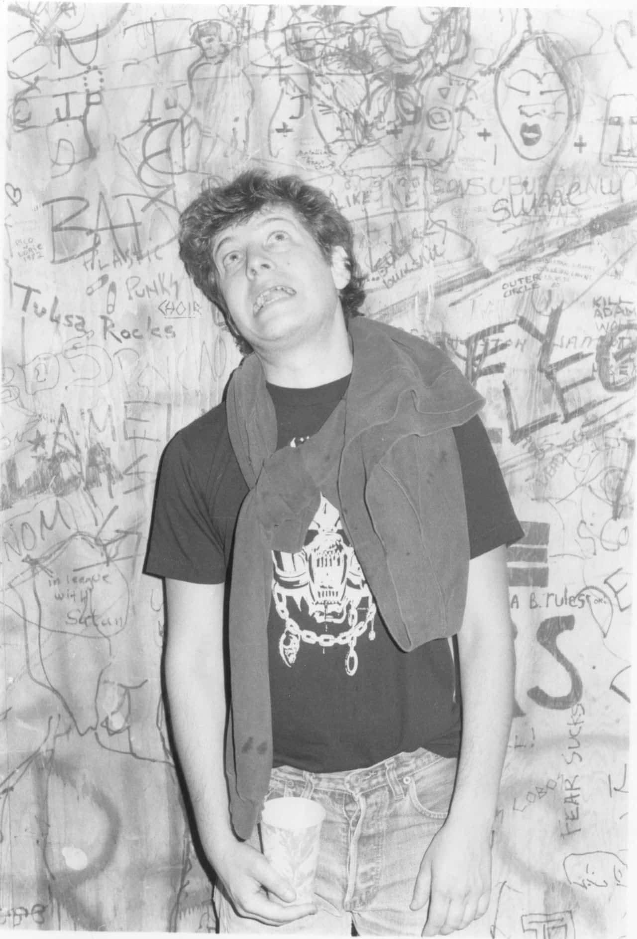 Black Flag Band 桌布 壁紙 — 一個穿著龐克T恤和外套的年輕男子站在一面佈滿混亂塗鴉和塗寫的牆前。