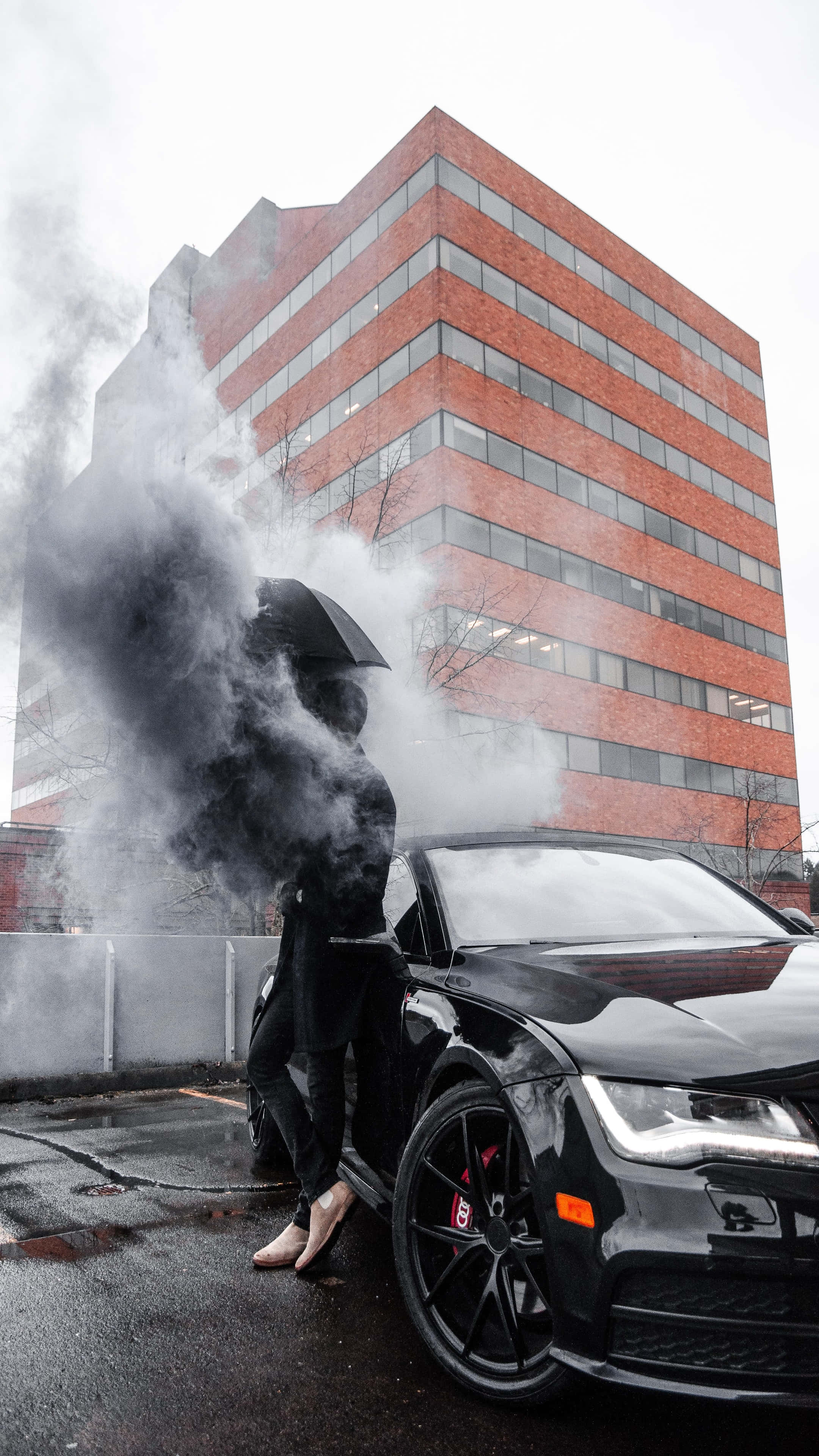 4K Car 桌布 壁紙 — 一個穿黑色衣服的人喺落雨嘅日子，企喺一個煙霧瀰漫嘅遮下面，喺一棟紅磚建築前面，企喺一架型格黑色跑車旁邊。