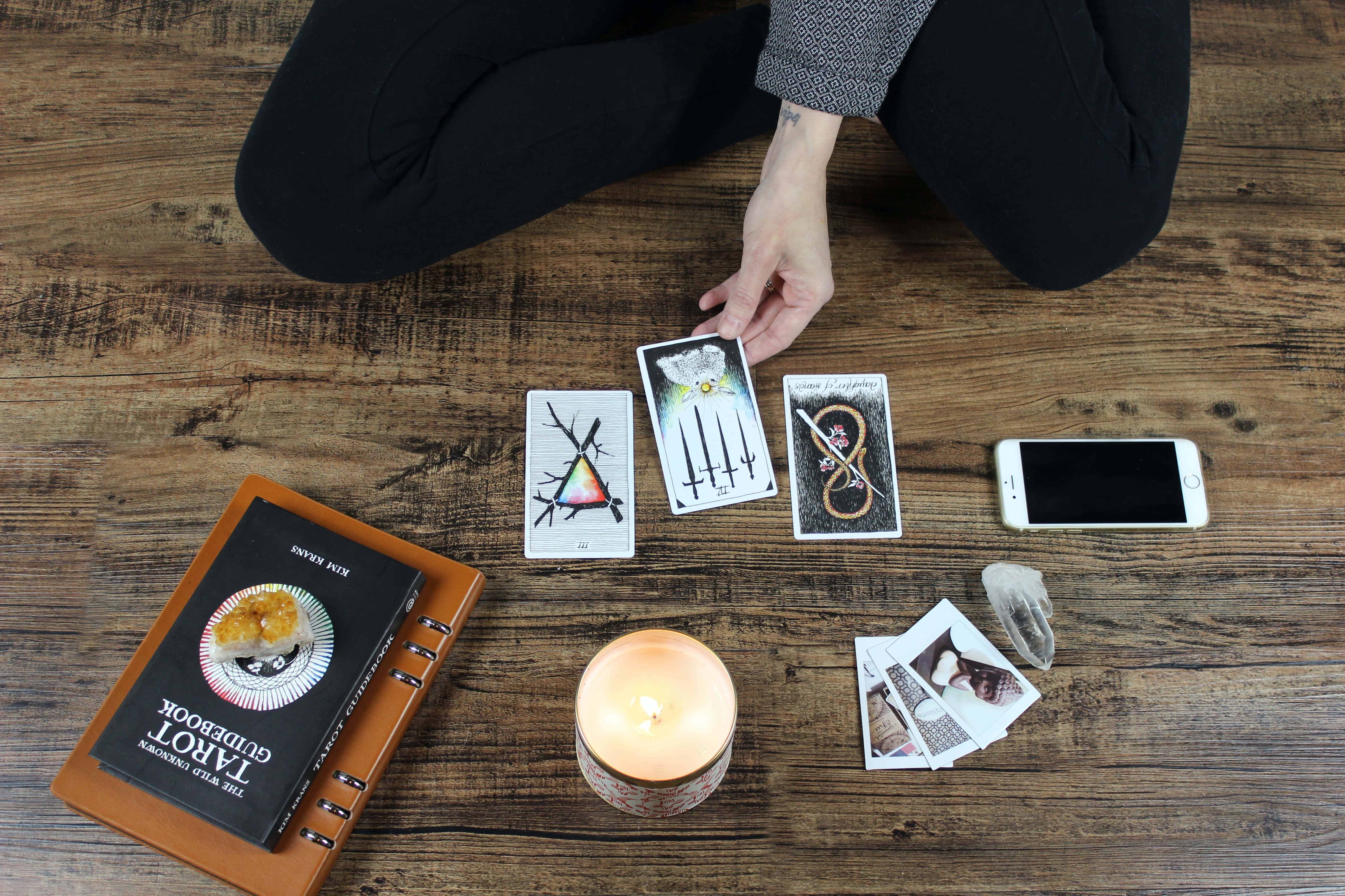 Tarot Card 桌布 壁紙 — 一個穿黑色褲子的人跪在質樸的木地板上，在點燃的蠟燭、塔羅指南書和智能手機旁邊排列塔羅牌。