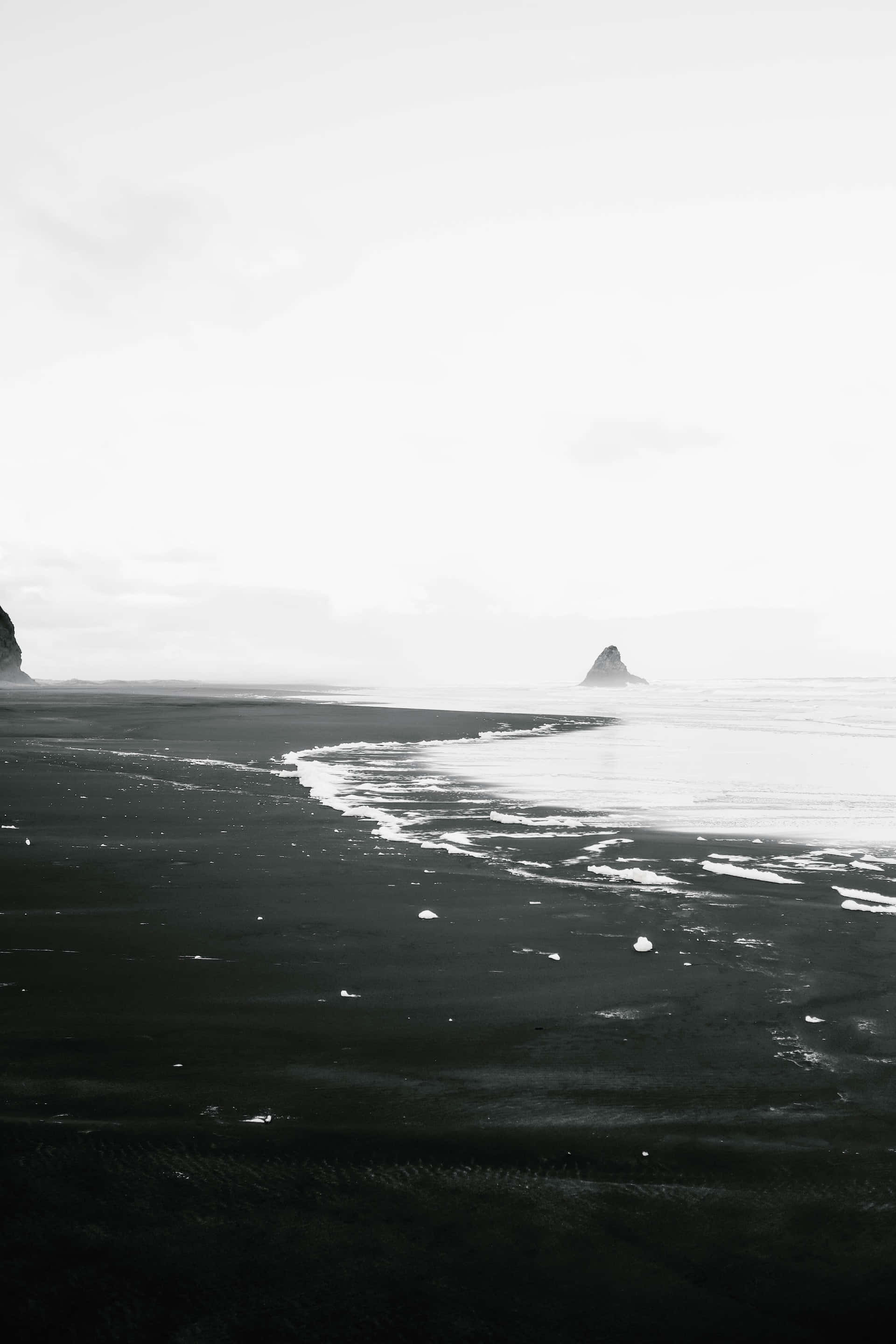 Black And White Beach 桌布 壁紙 — 一個突出的黑沙海灘在朦朧多雲的天空下與平靜的海浪和遠處的岩石形成相遇。